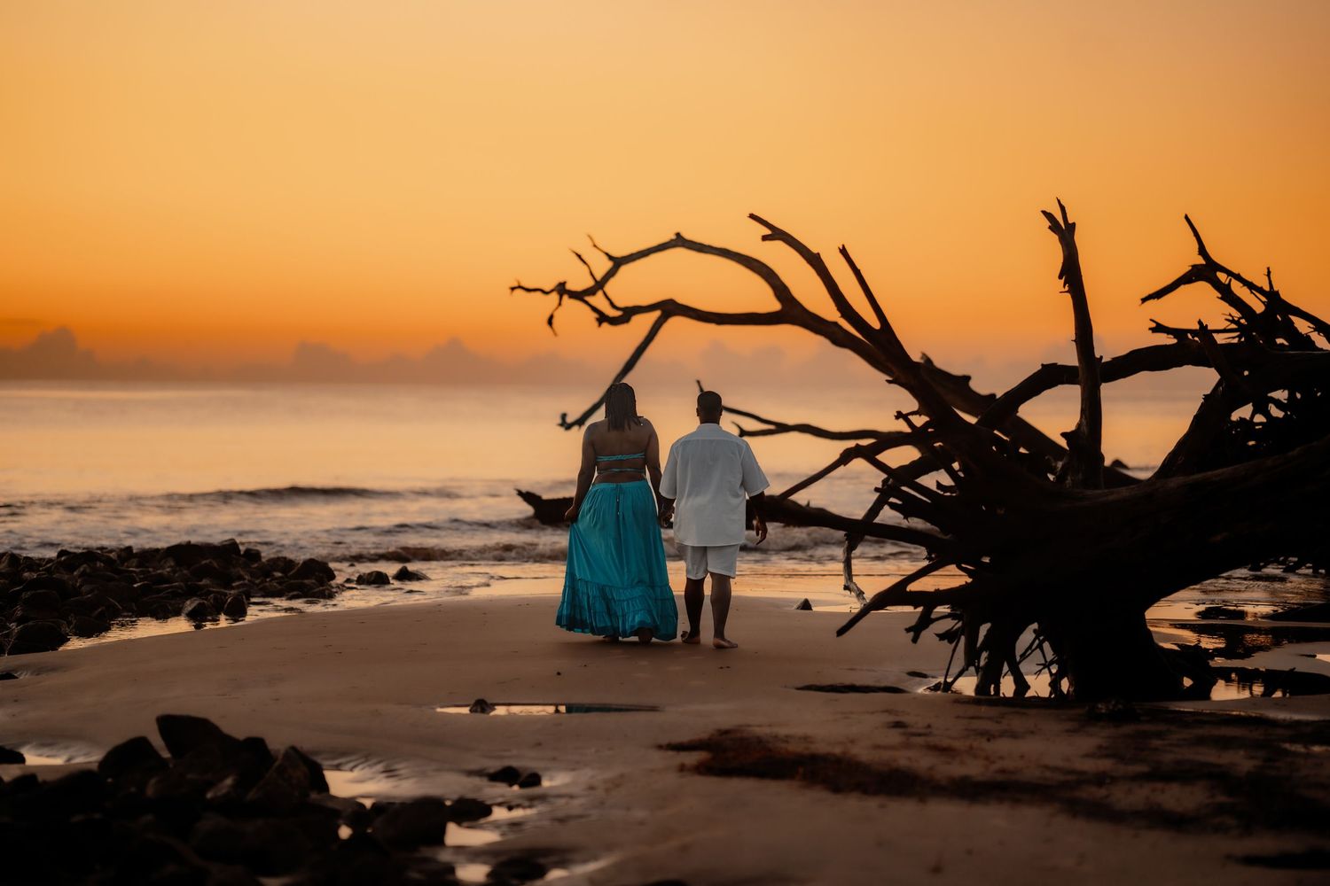 Couple standing on beach at golden sunset near driftwood by ocean waves.
