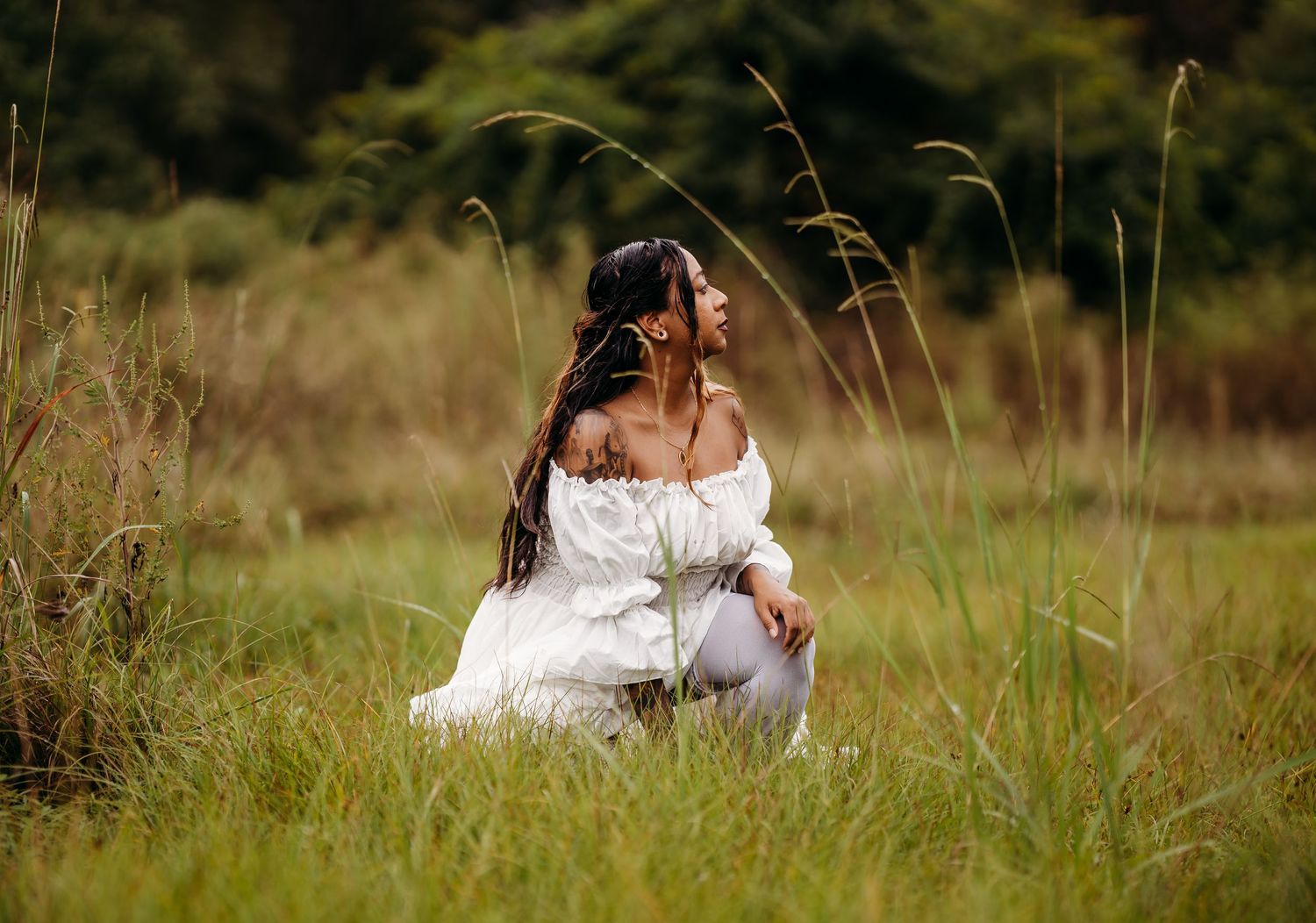 Woman in white off-shoulder dress crouching in a lush green meadow with tall grass.