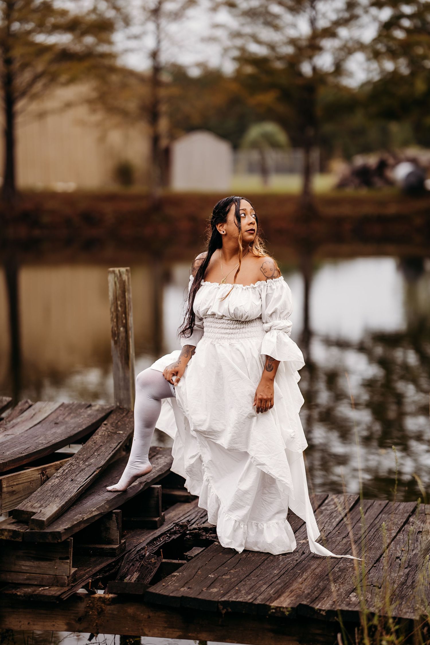 Woman in white ruffled off-shoulder gown sits elegantly on old wooden dock by reflective lake.