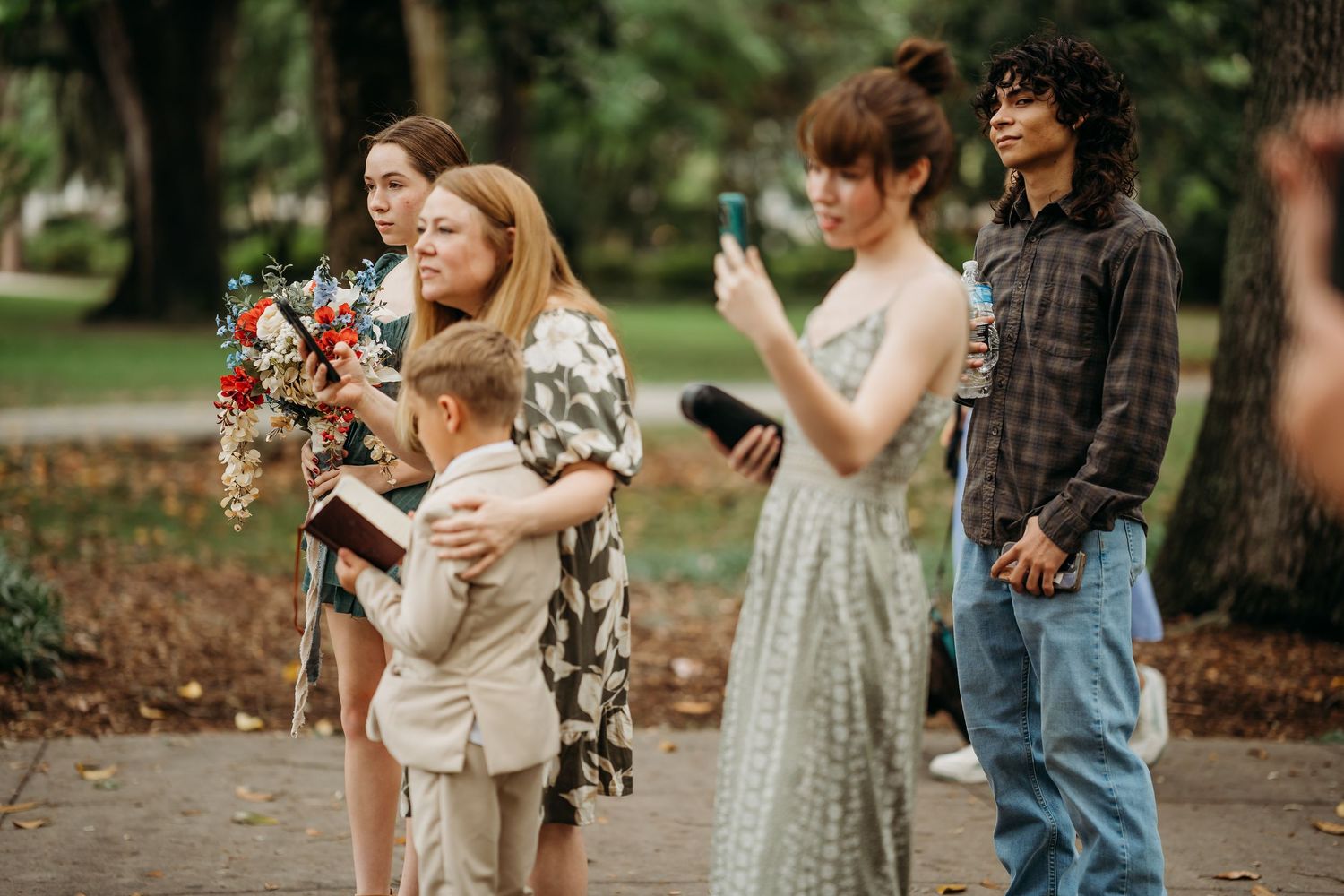 Wedding guests including a young boy and woman with bouquet watch an outdoor park wedding ceremony.