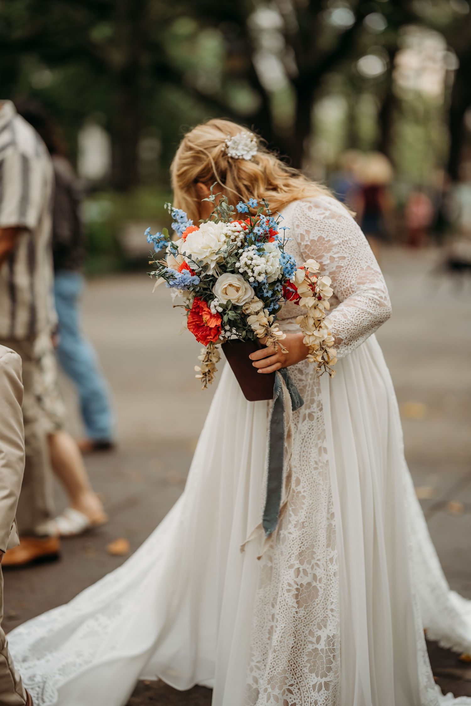 Bride in lace wedding dress smelling colorful bouquet of red, white, and blue flowers.
