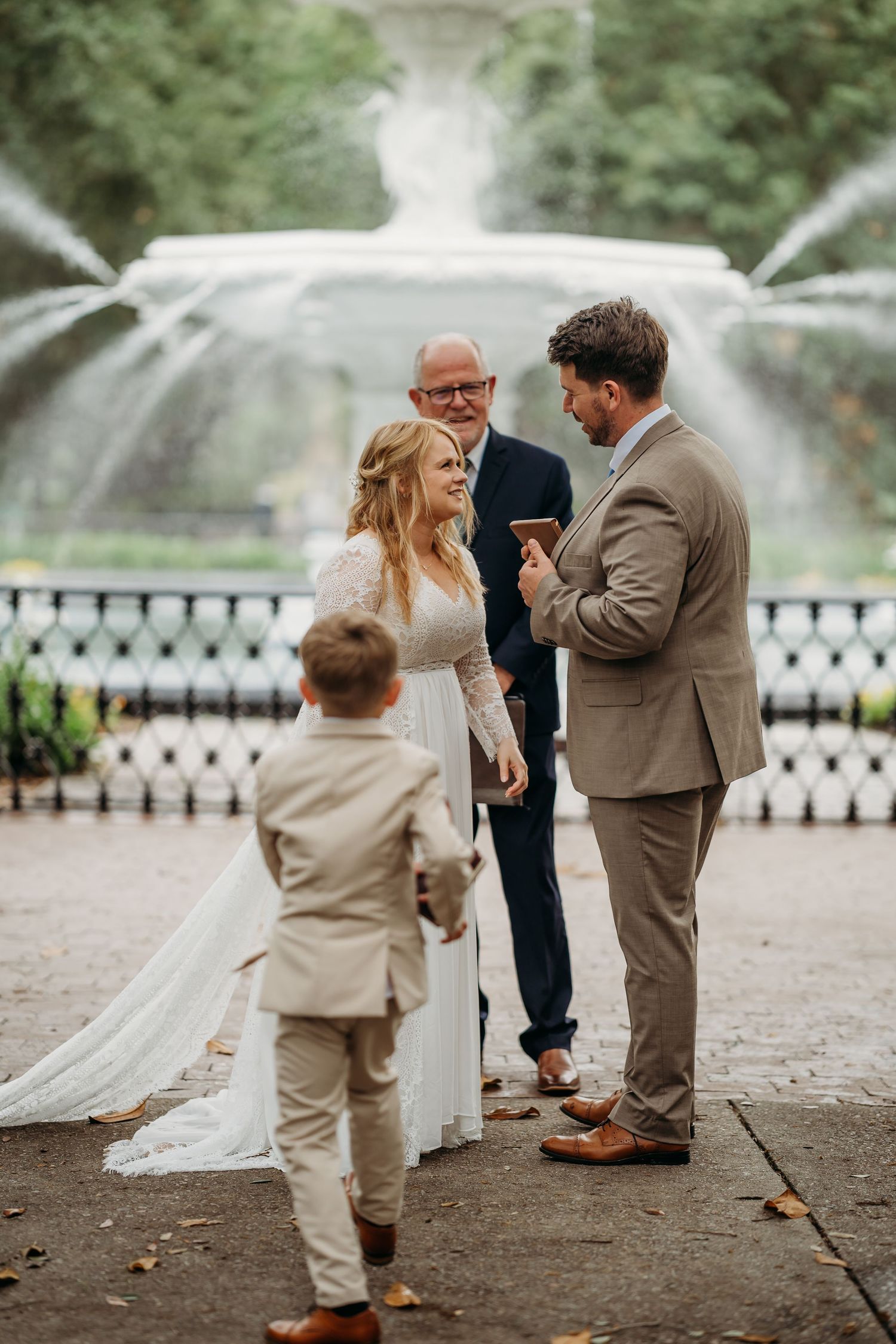 Couple exchanges vows with officiant at elegant outdoor wedding ceremony in front of large fountain.