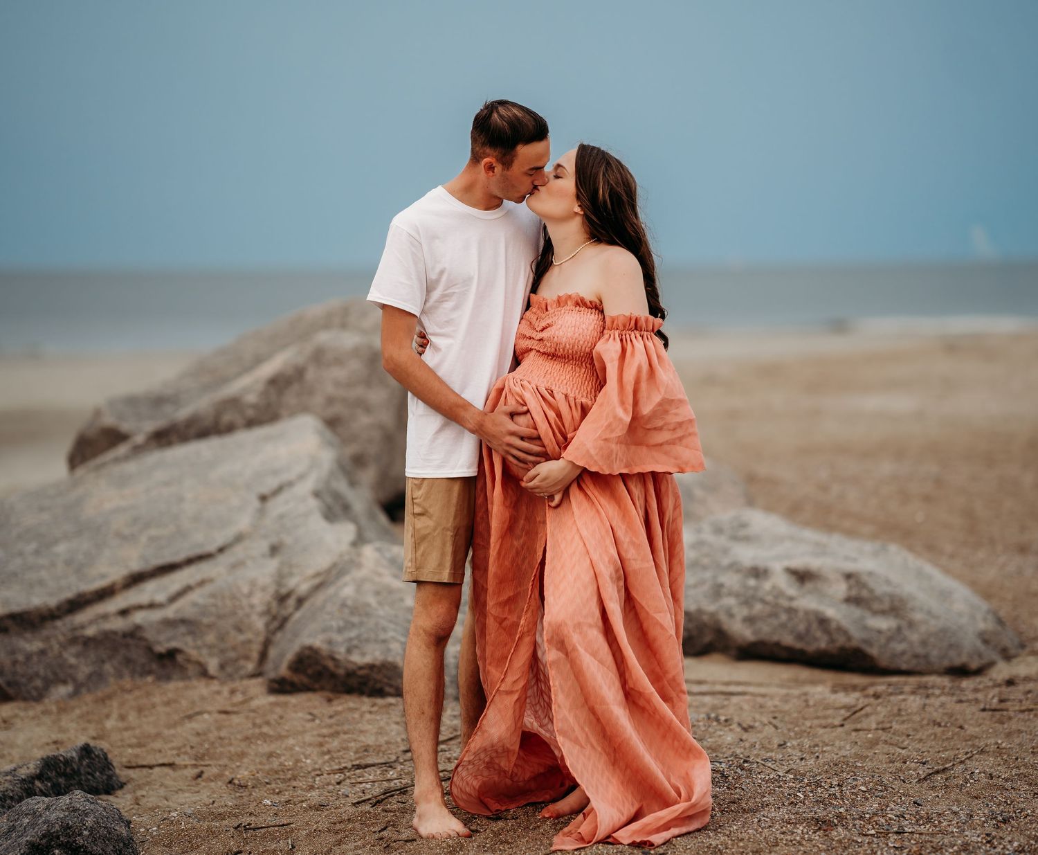 Pregnant couple sharing a kiss on a rocky beach, woman in coral off-shoulder maternity gown.