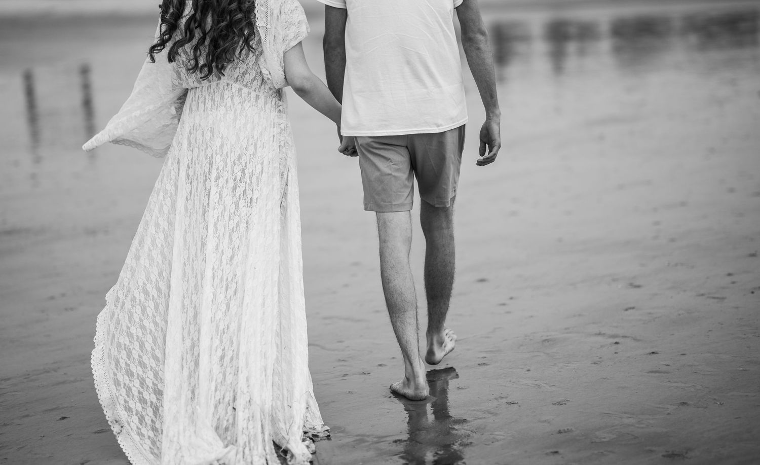 Black and white photo of pregnant couple walking barefoot hand in hand along a reflective beach shoreline.