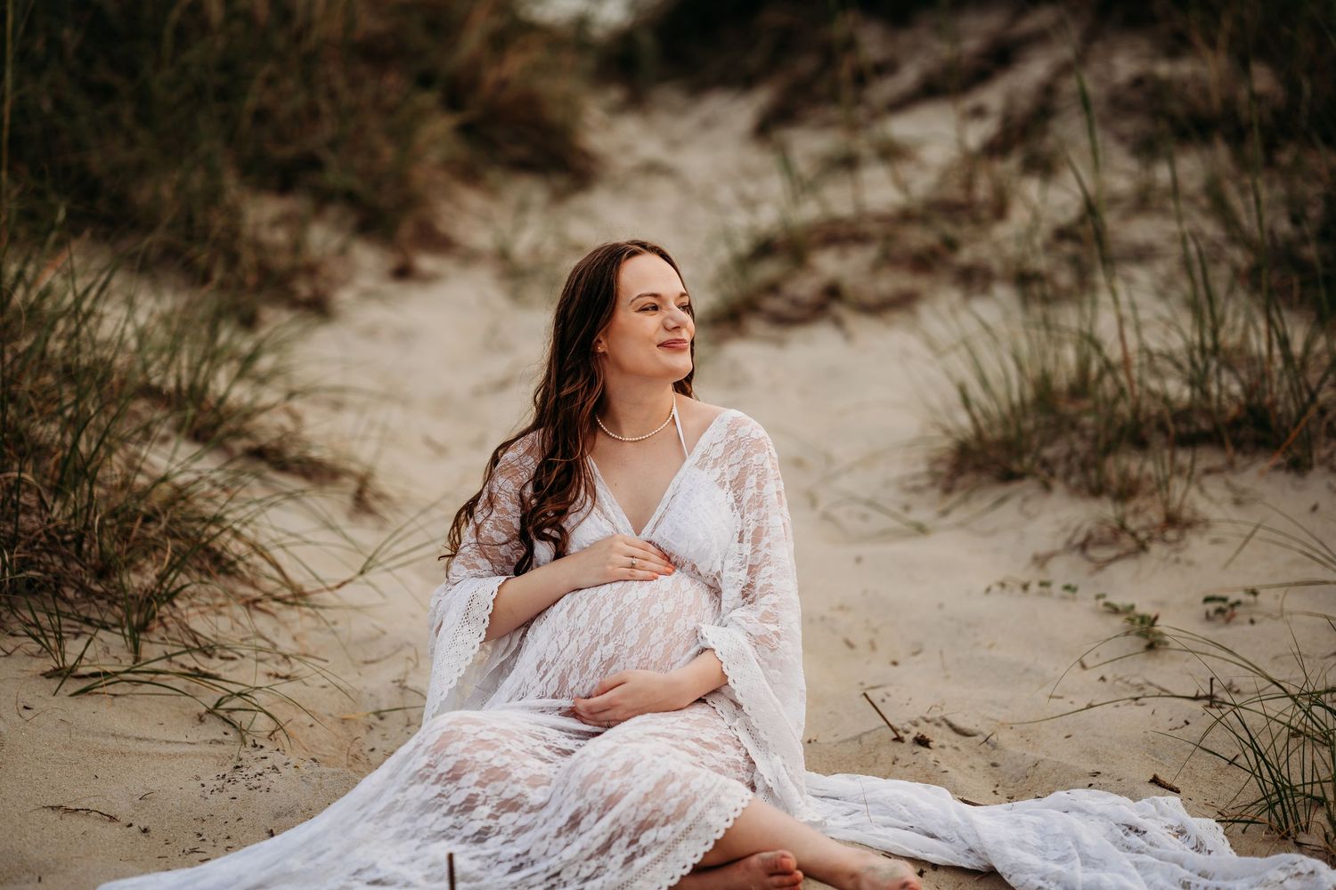 Pregnant woman in white lace dress sitting in sand dunes, smiling softly during golden hour.
