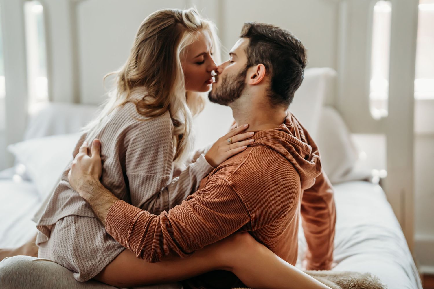 Affectionate couple kissing while sitting on a bed in a warmly lit bedroom.