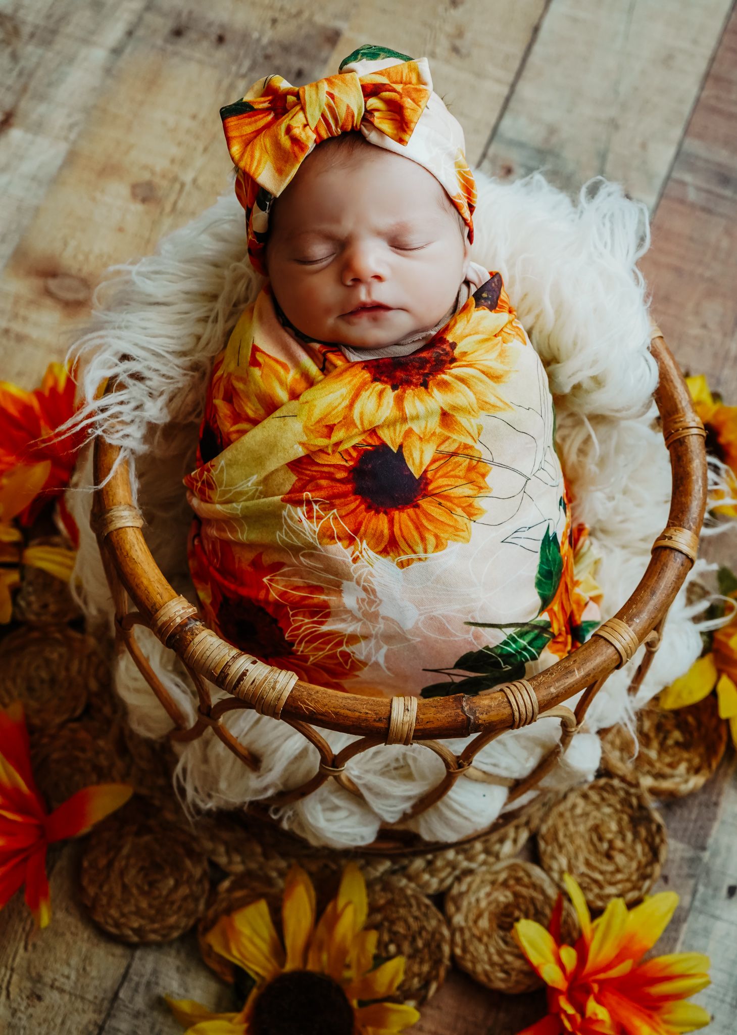 Sleeping newborn wrapped in sunflower swaddle resting in wicker basket with flowers.