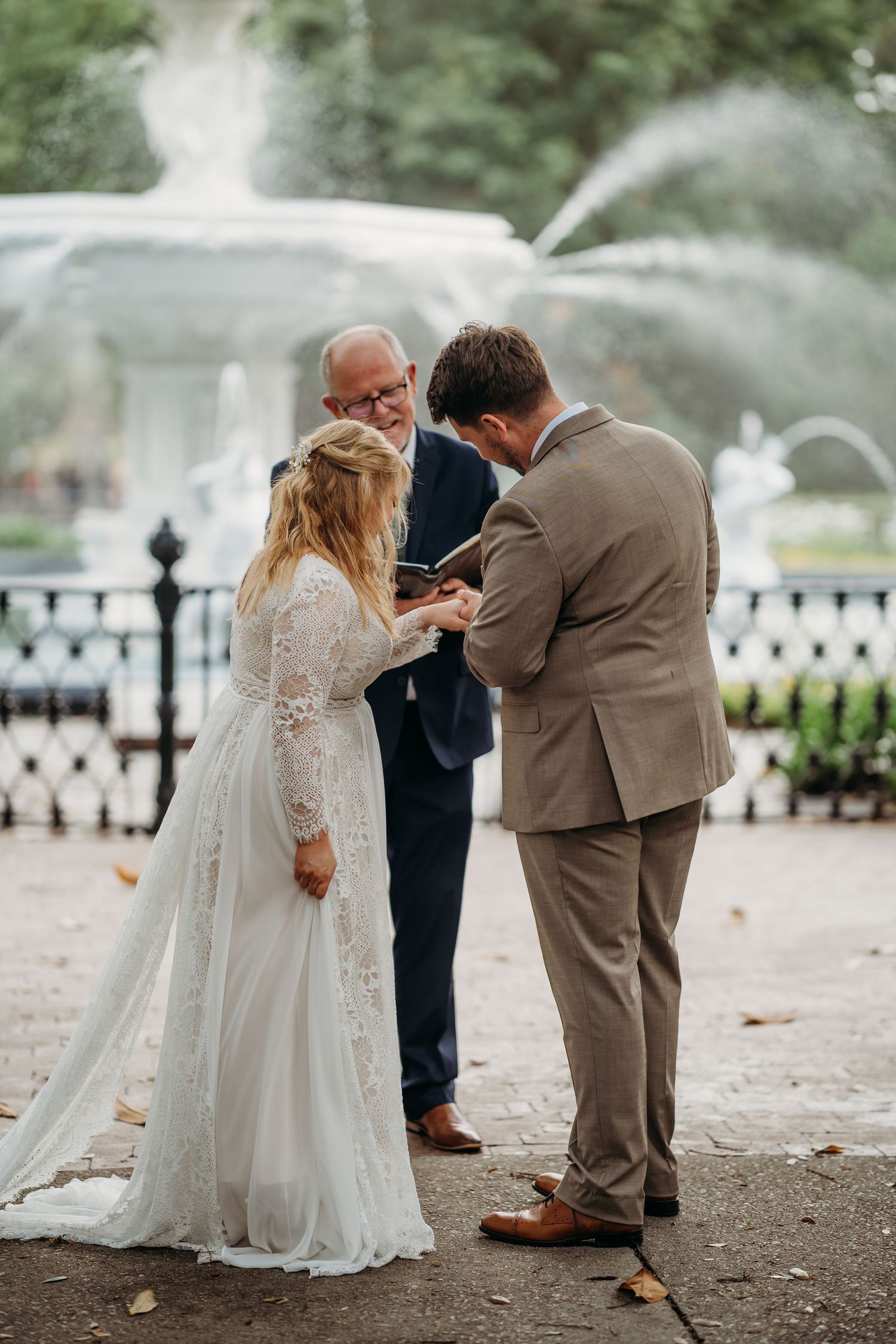 Groom places wedding ring on bride's finger as smiling officiant watches at fountain ceremony.