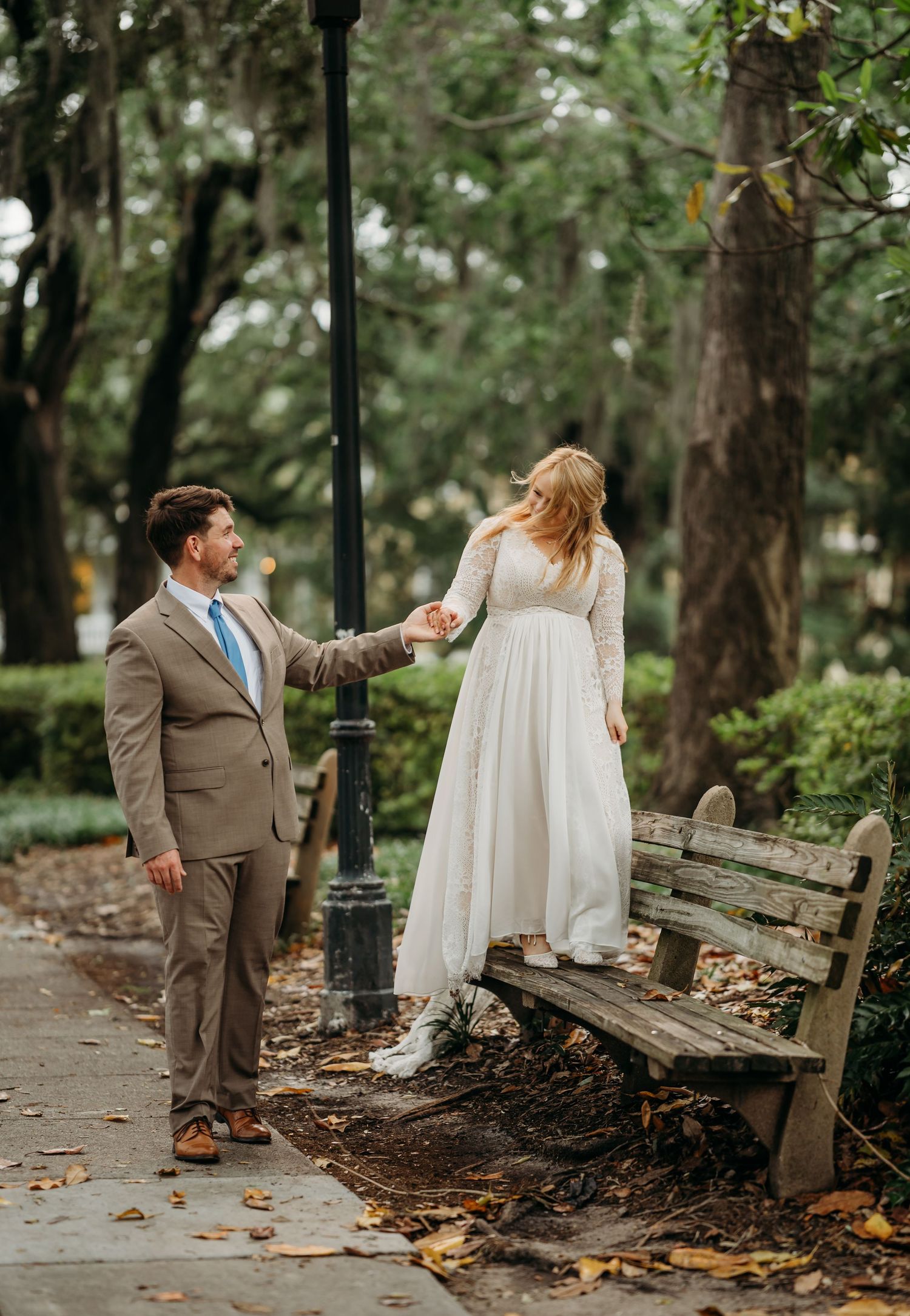 Groom helps bride step down from a park bench while holding hands in a lush garden setting.