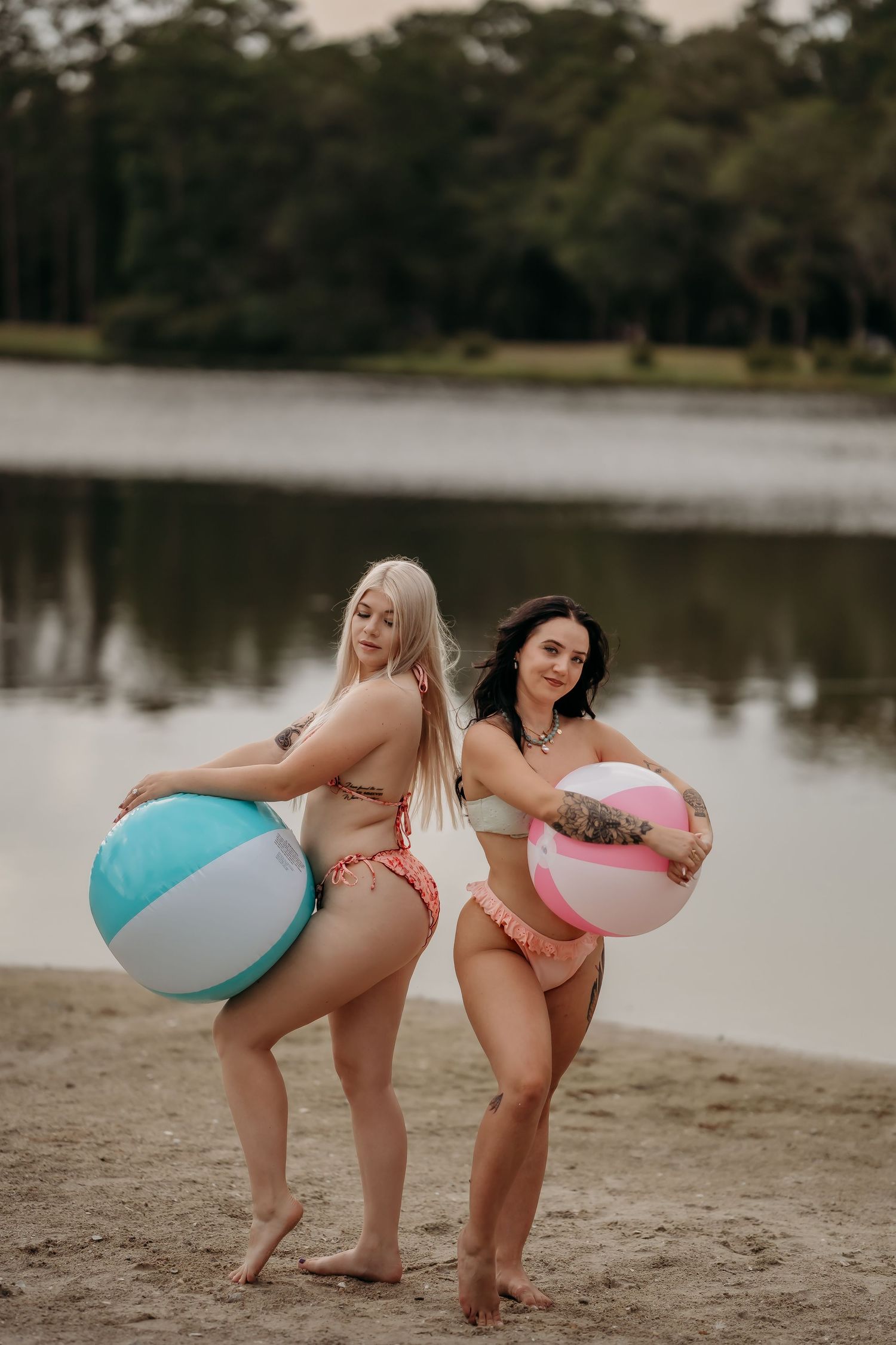 Two women in bikinis pose back-to-back holding colorful beach balls on a sandy lakeside beach.