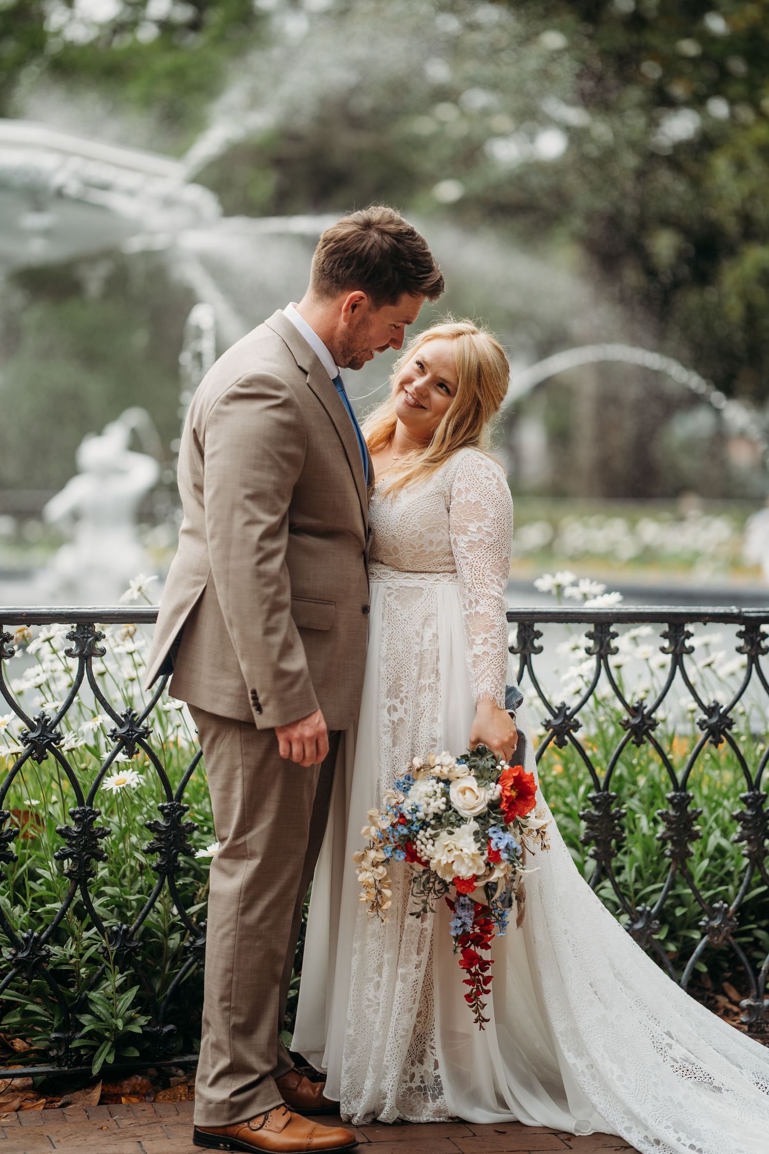 Bride and groom share a tender moment in front of a fountain, she holds a colorful bouquet in a lace wedding gown.