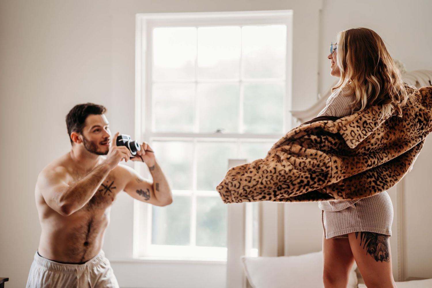 Shirtless man photographing woman playfully swinging leopard coat in bright bedroom.