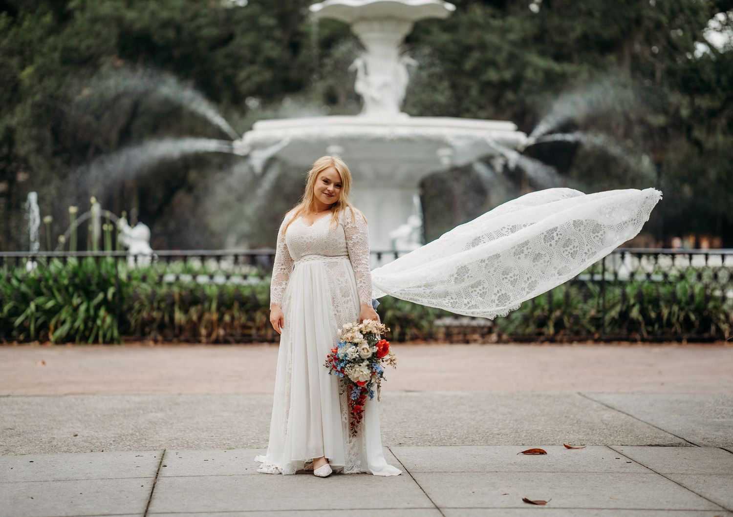 Plus-size bride in long-sleeve lace wedding dress holds colorful bouquet as veil flows in breeze by fountain.