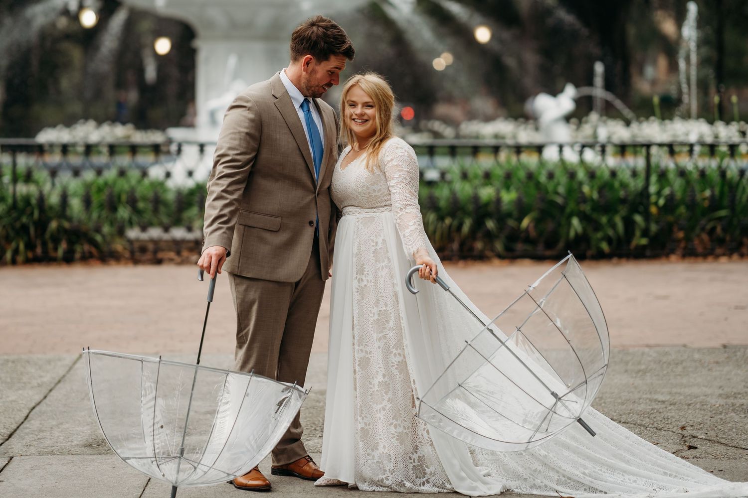 Groom in tan suit gazes lovingly at smiling bride in lace gown holding clear umbrellas by fountain.