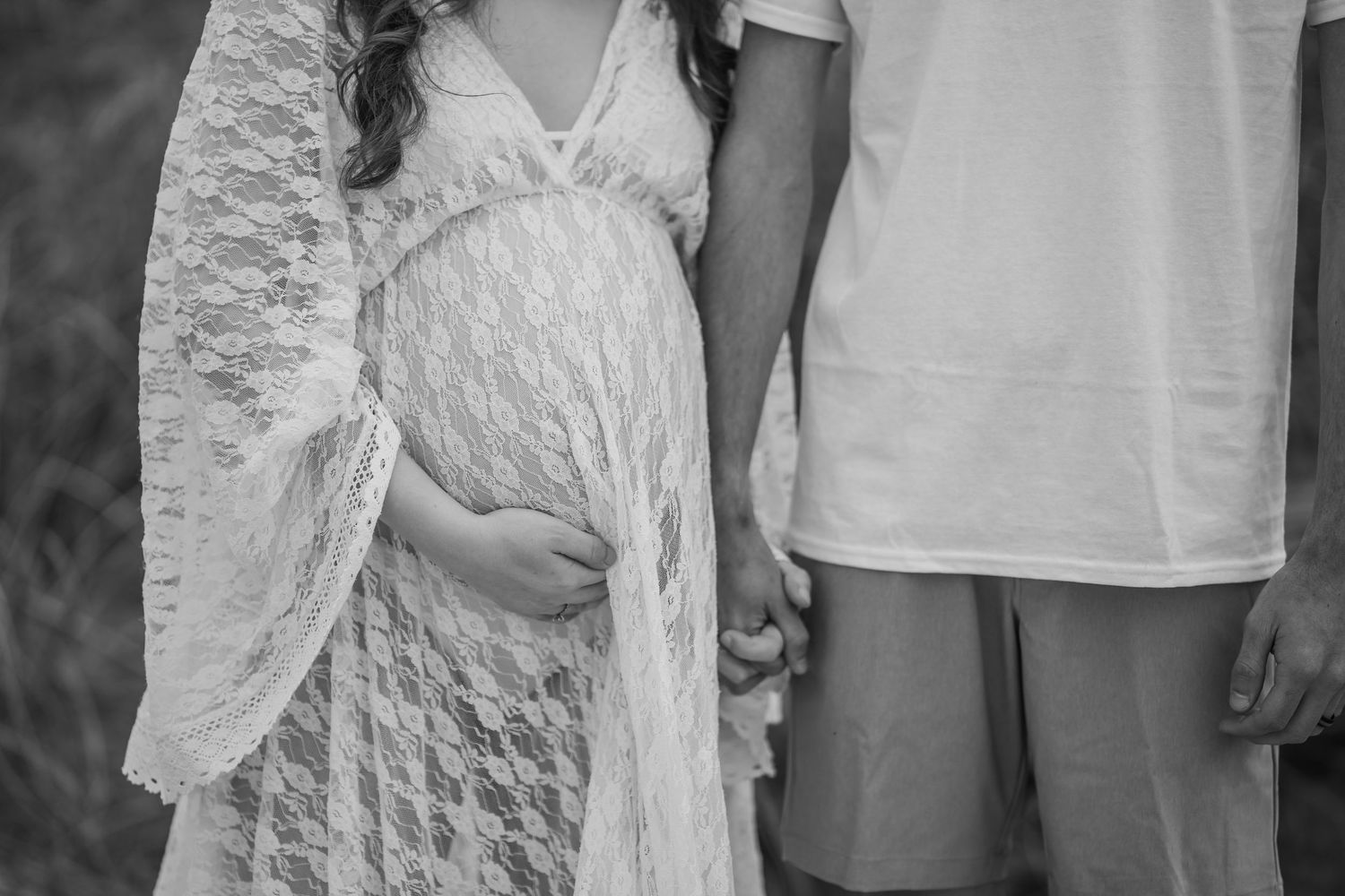 Black and white close-up of pregnant woman in lace dress holding baby bump with partner beside her.