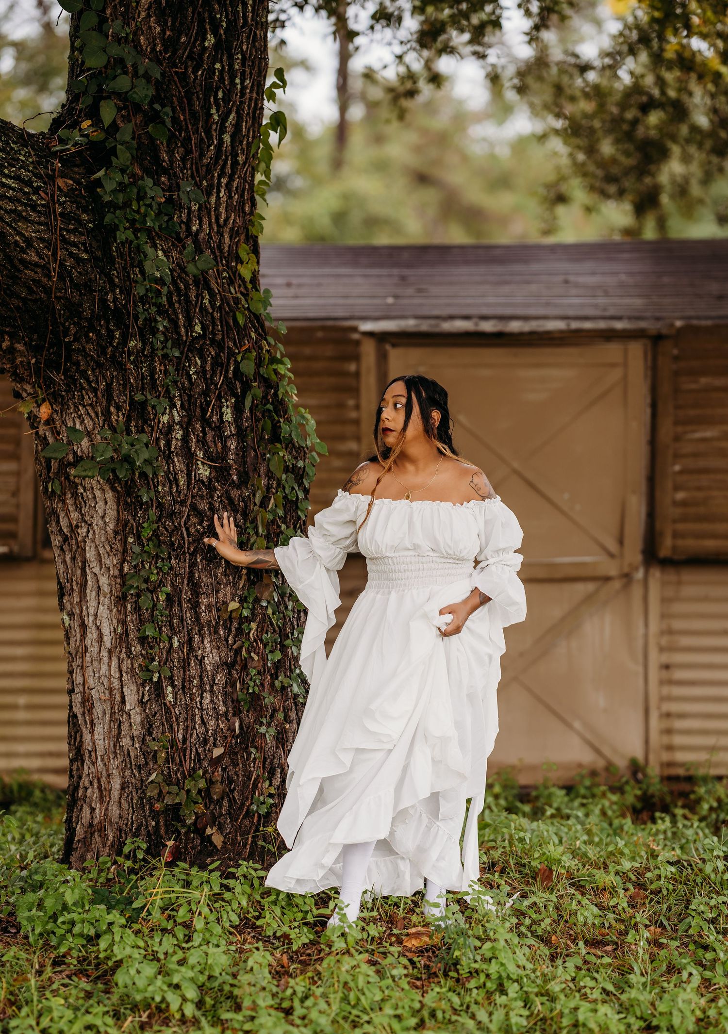 Woman in white off-shoulder dress posing by a large tree near a rustic wooden shed outdoors.