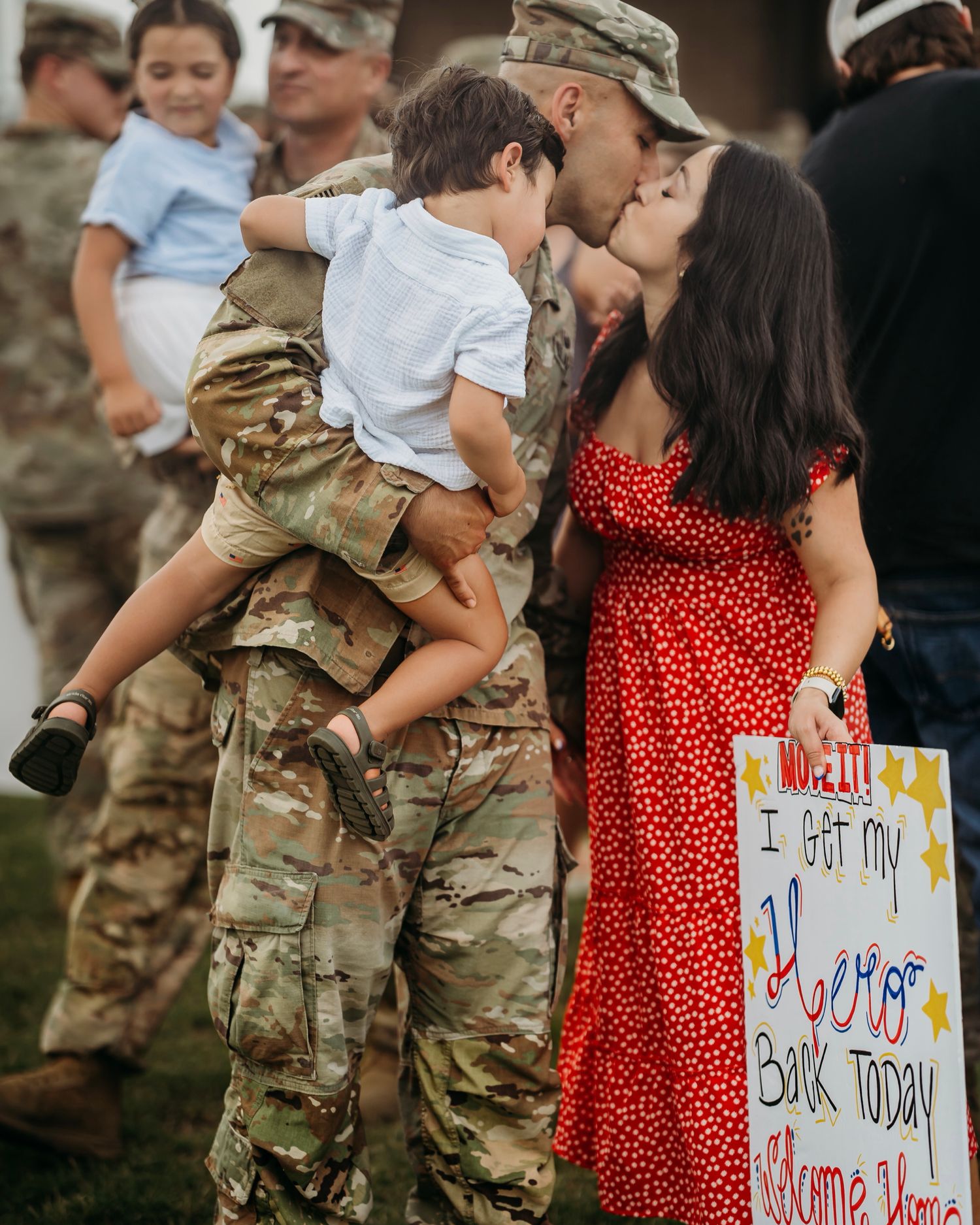 Soldier in camouflage kisses wife in red dress at homecoming while holding children, with welcome home hero sign.