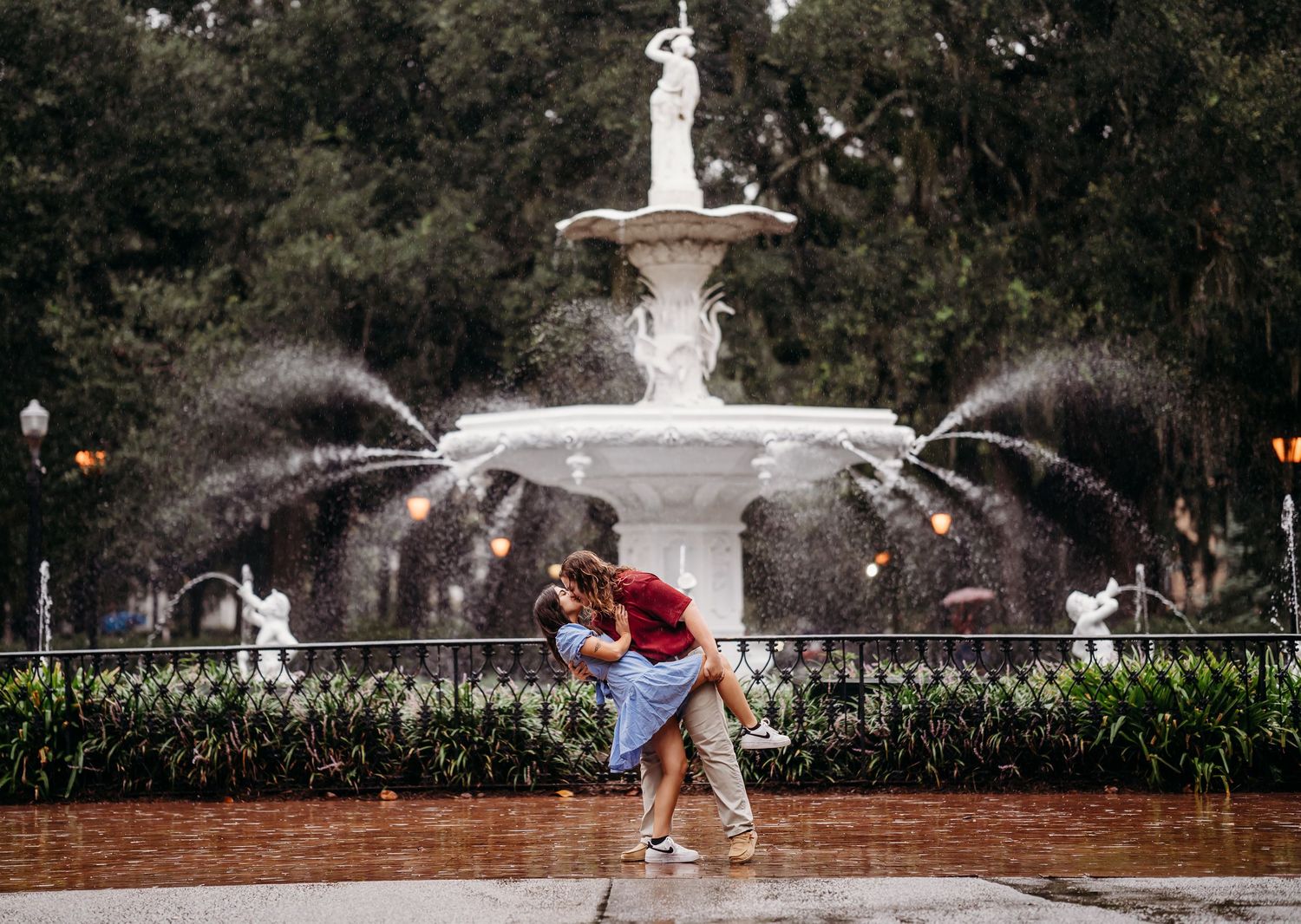 Couple dancing romantically in the rain in front of a white ornate fountain in a lush green park.