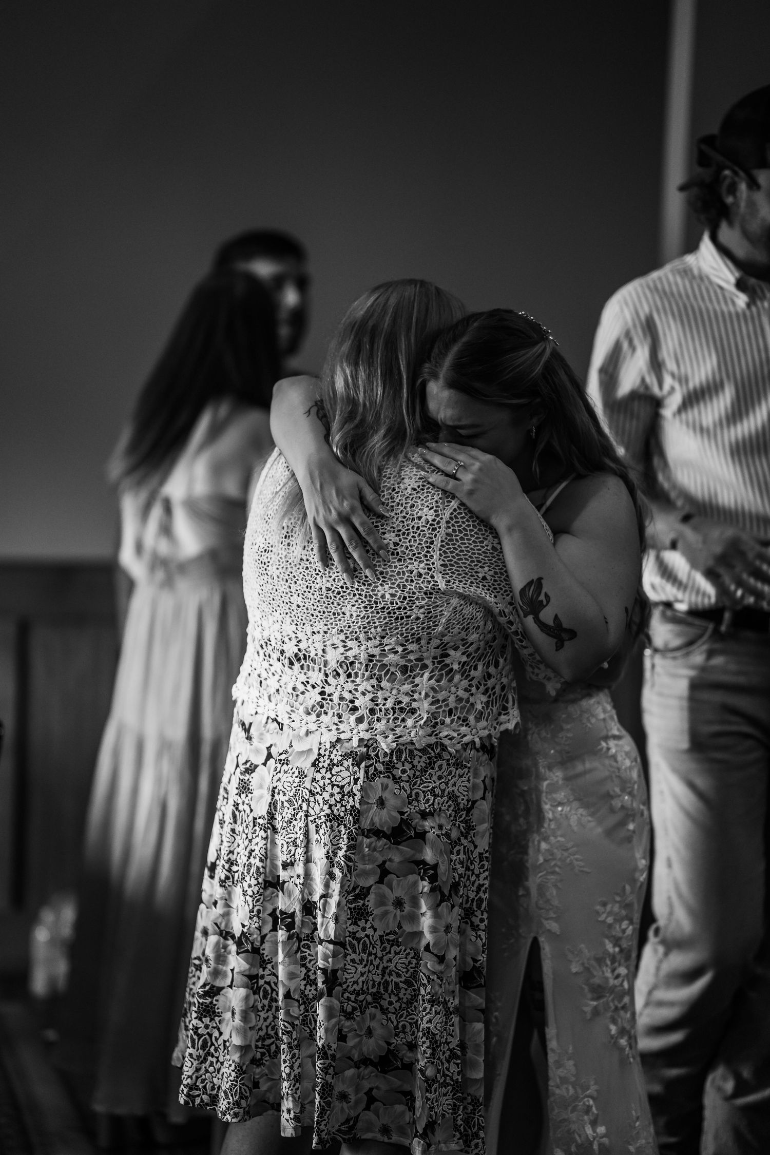 Black and white photo of two women sharing an emotional hug at a social gathering, with guests visible in background.