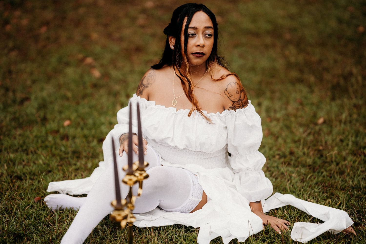Woman in white off-shoulder dress sitting on grass with candelabra nearby.