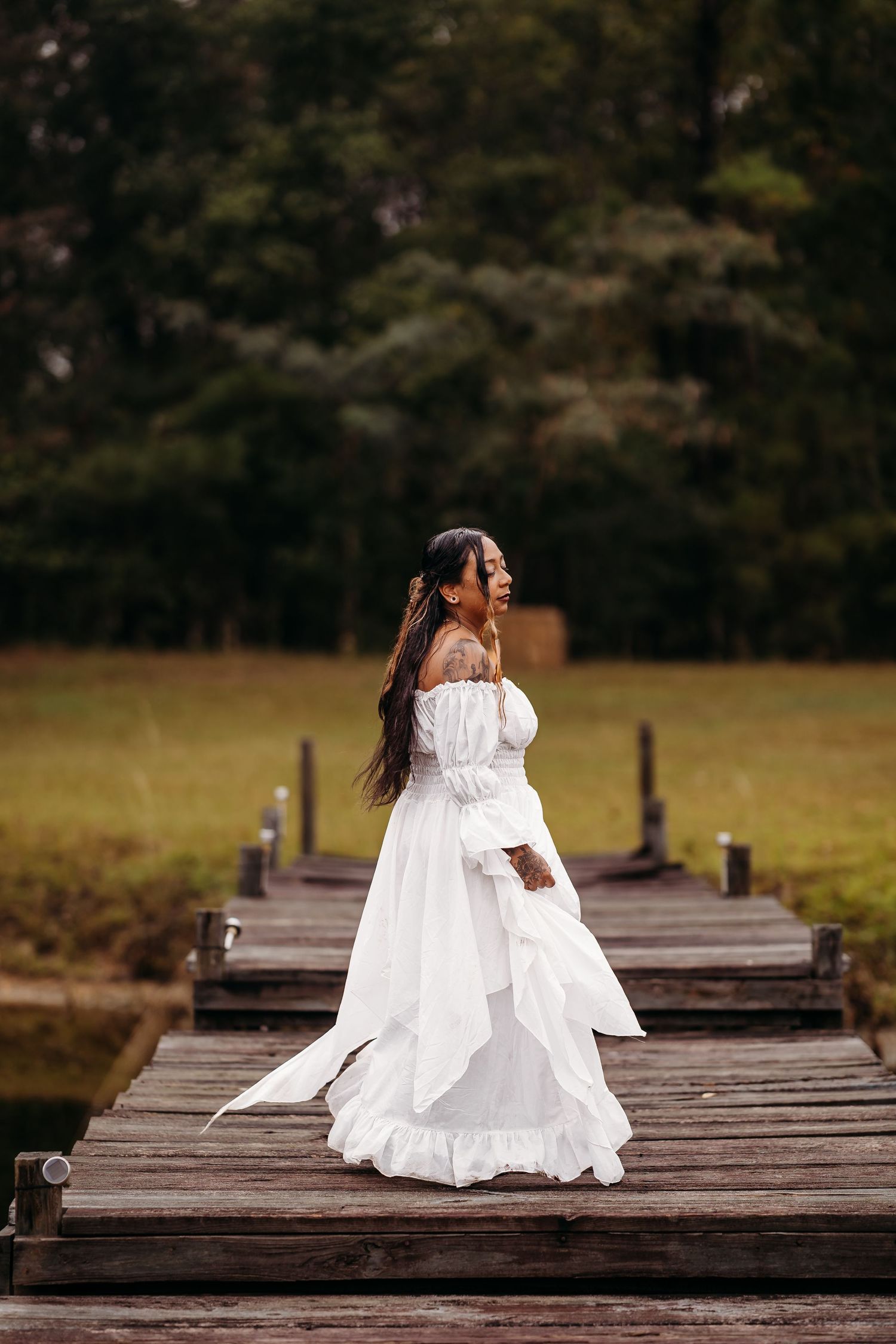 Bride in white off-shoulder ruffled gown stands on a wooden dock surrounded by lush greenery.
