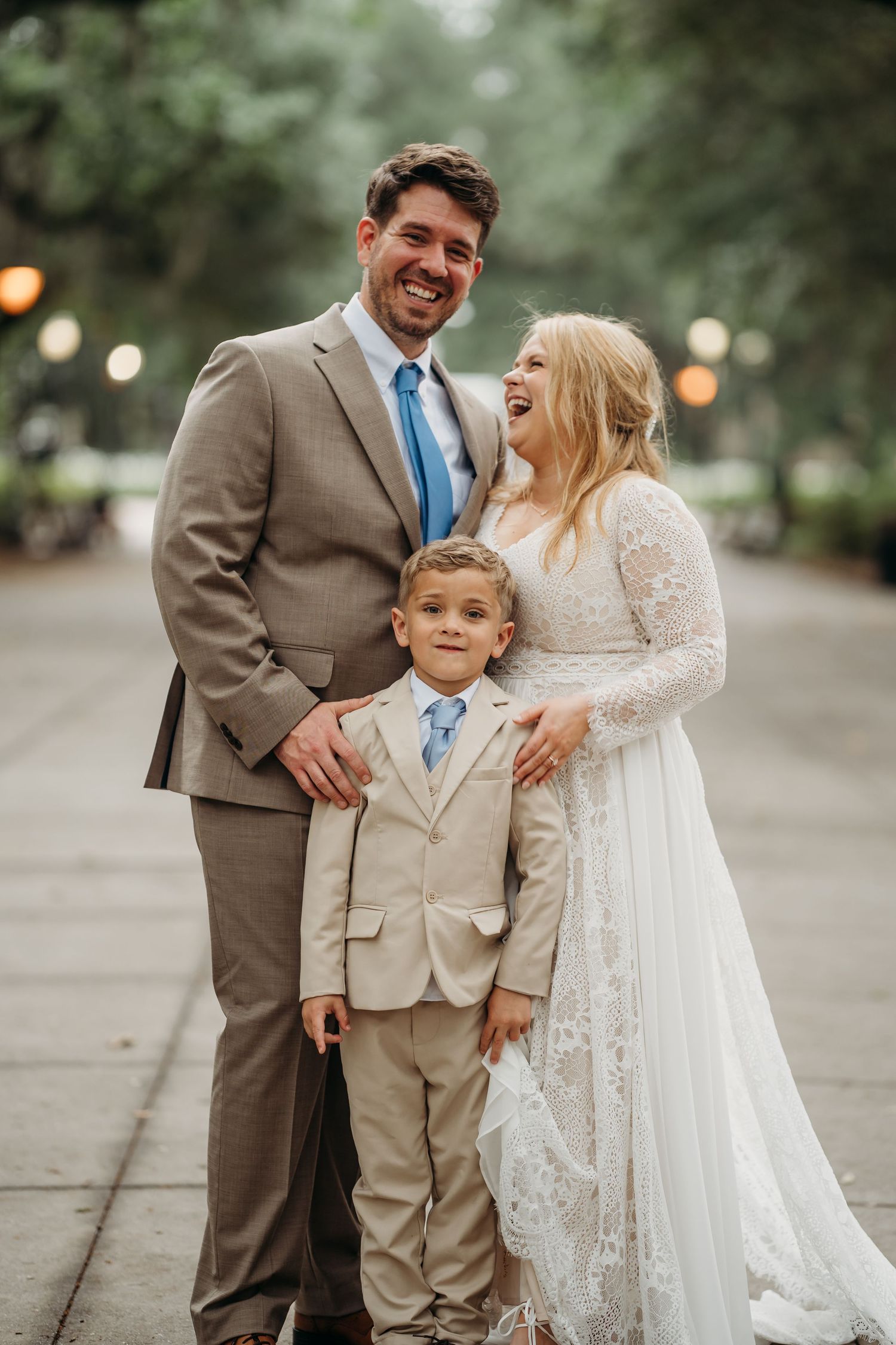 Happy wedding couple and young boy in blue tie pose together on Forsyth Park pathway in Savannah.