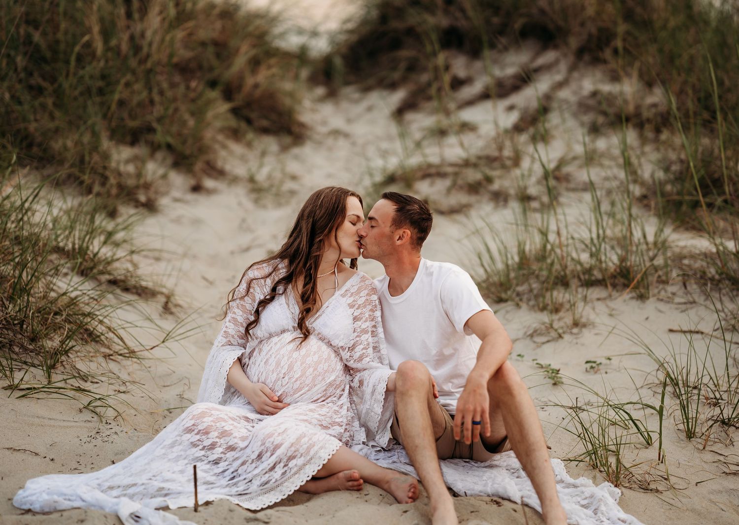Pregnant couple kissing while seated on sandy beach dunes, woman in white lace maternity dress.