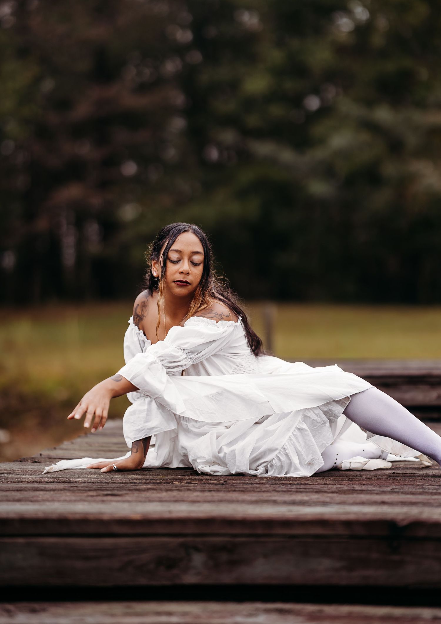 Elegant woman in white off-shoulder gown poses gracefully seated on a rustic wooden dock.