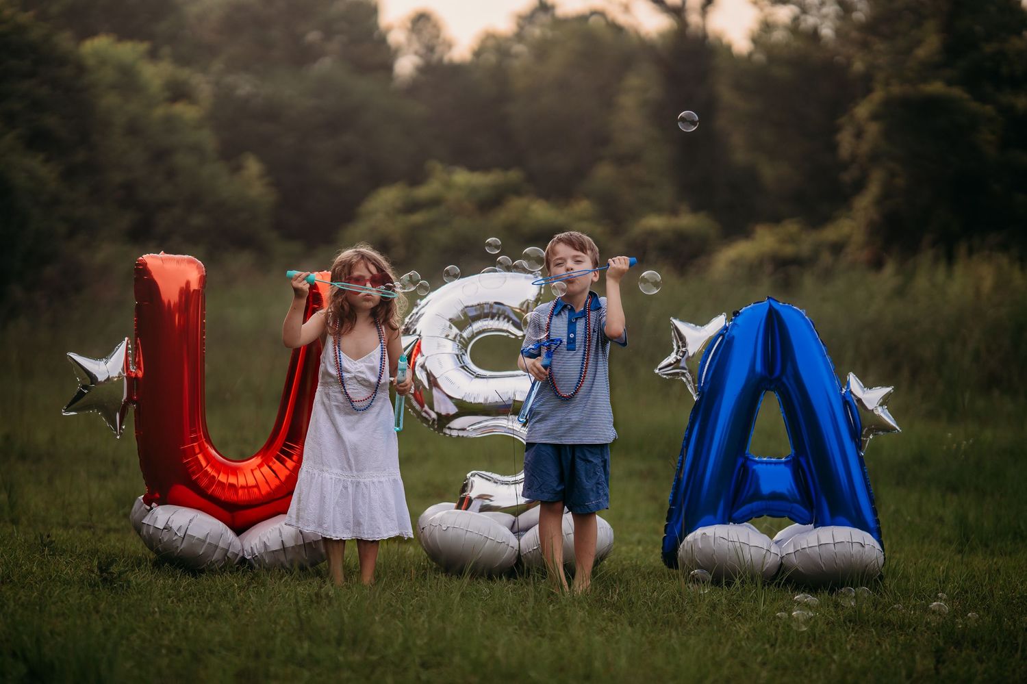Two children pose with red, silver, and blue USA letter balloons on grass, blowing bubbles, celebrating Fourth of July.