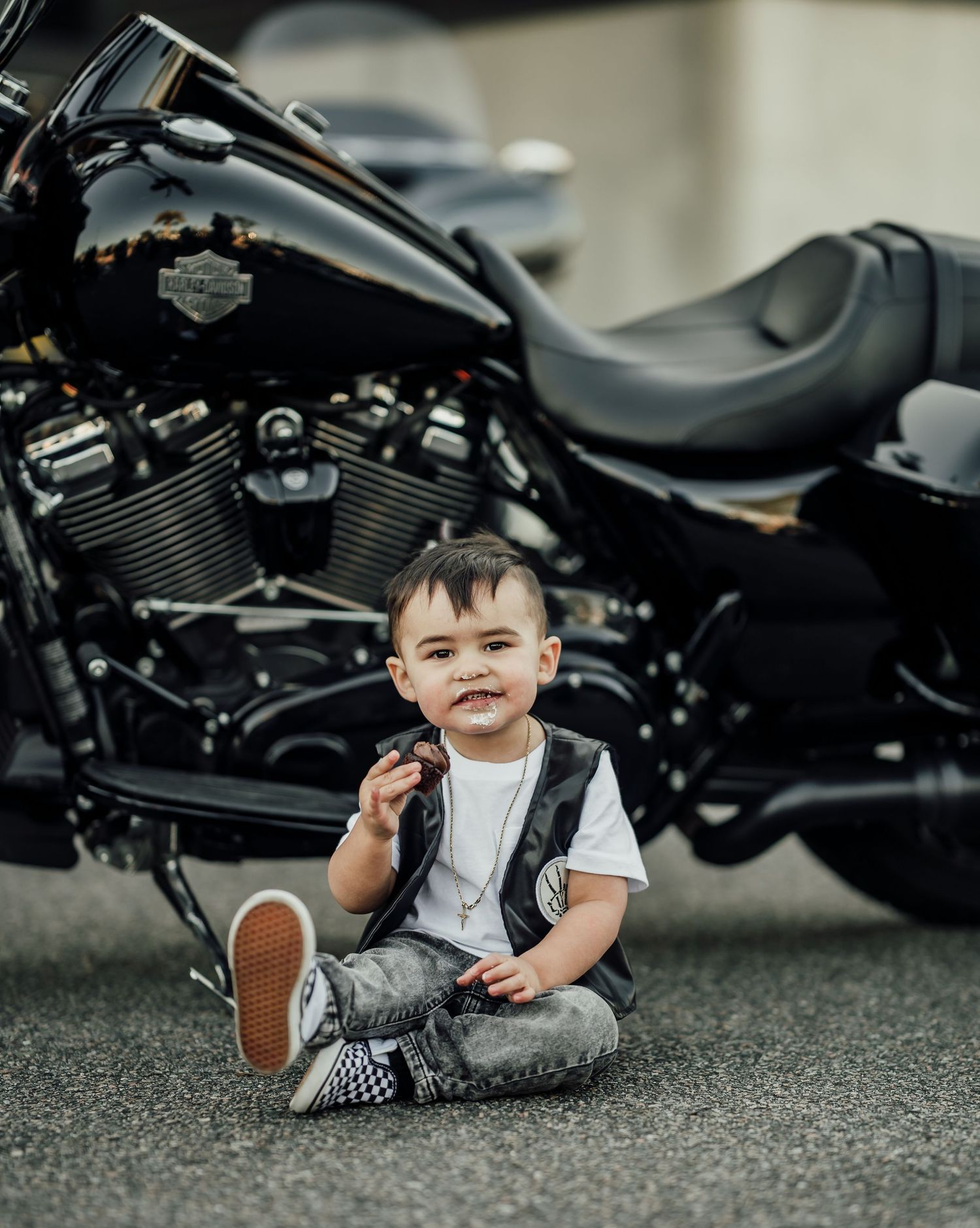 Smiling baby boy in leather vest and jeans sitting on pavement in front of a black Harley-Davidson motorcycle.