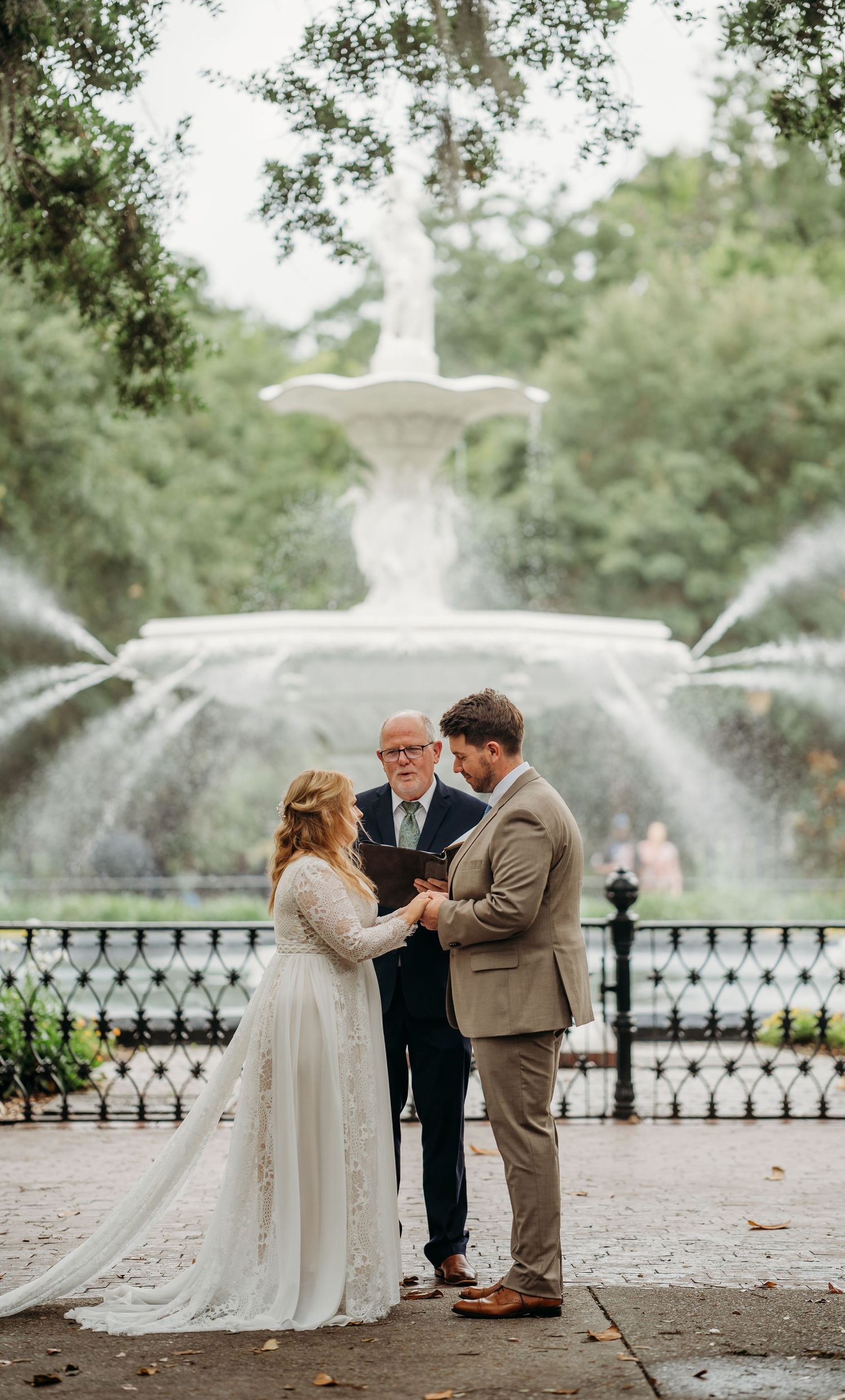 Bride and groom exchange vows at Forsyth Park fountain in Savannah, GA with officiant during outdoor wedding ceremony.