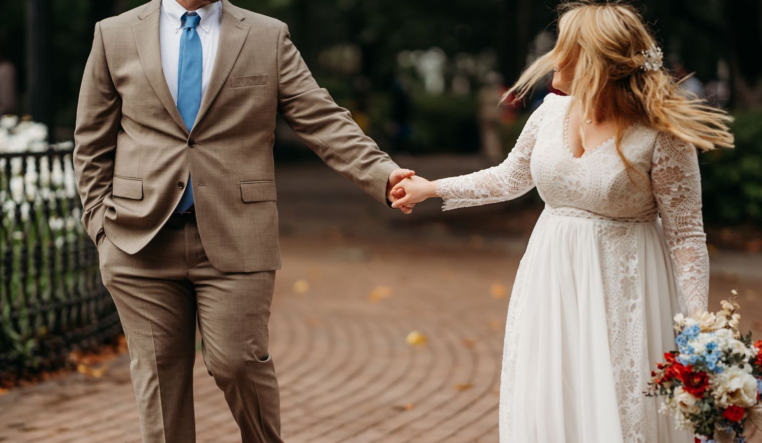 Bride and groom walk hand in hand on a brick path, bride carrying a vibrant red and blue bouquet in lace gown.