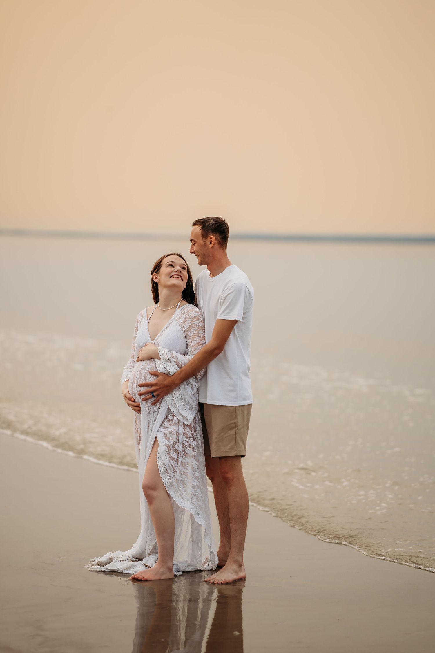 Pregnant couple embracing on beach at sunset, woman in white lace dress cradling baby bump.