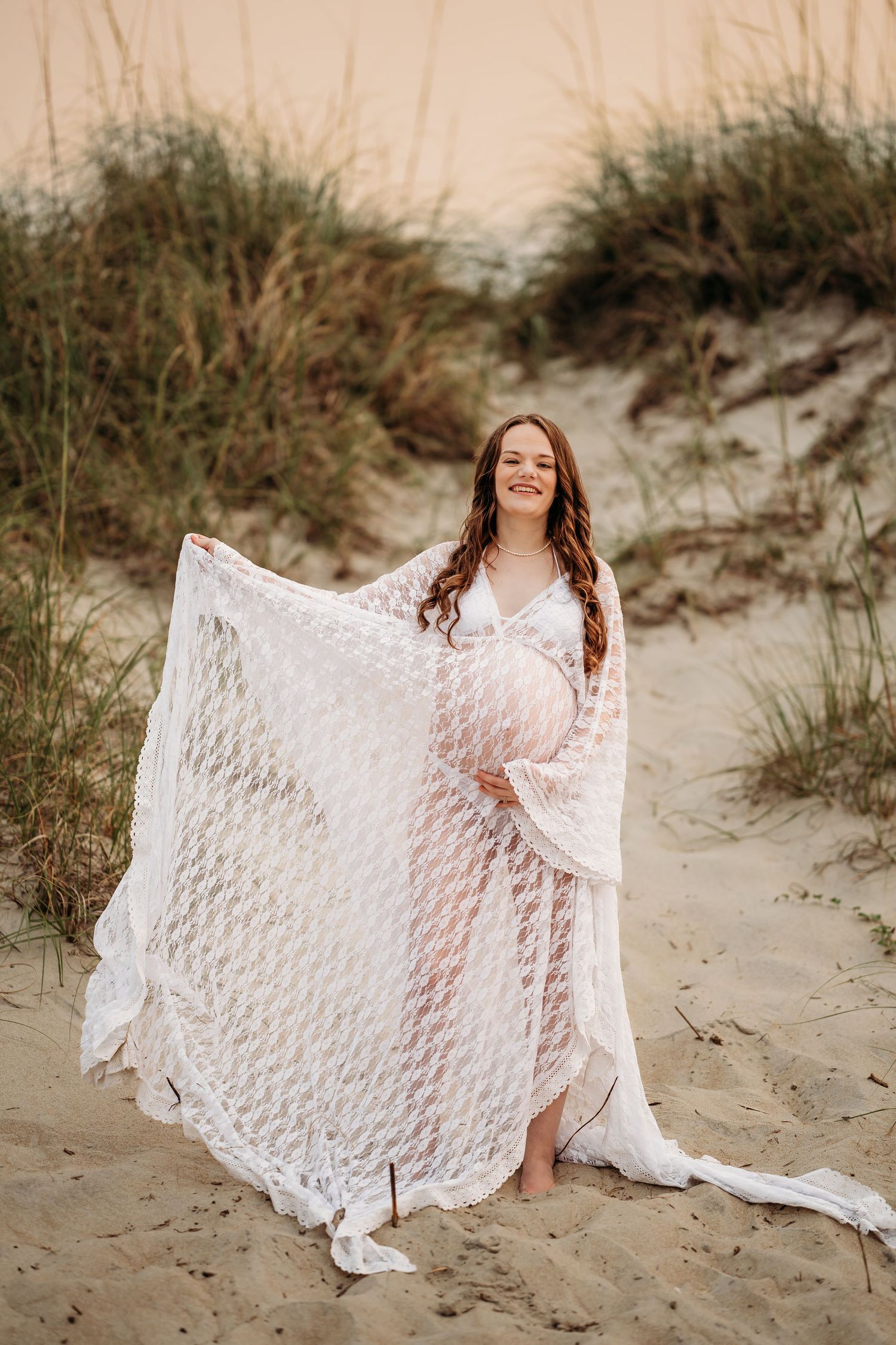 Joyful pregnant woman in white lace maternity dress posing on sandy beach dunes at sunset.