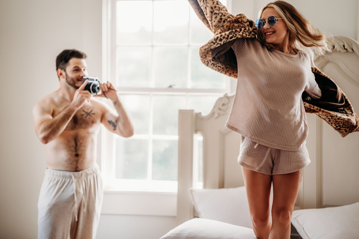 Tattooed man with camera photographing woman in sunglasses swinging leopard coat on bed.
