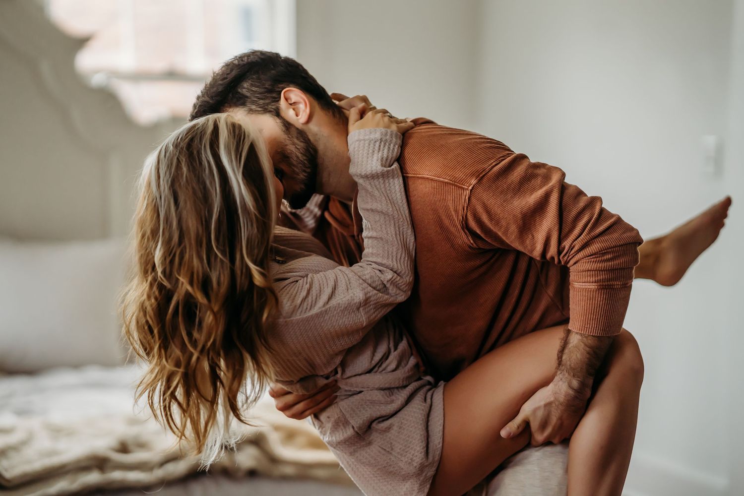 A man playfully dips a laughing woman on a bed in a bright, cozy bedroom setting.