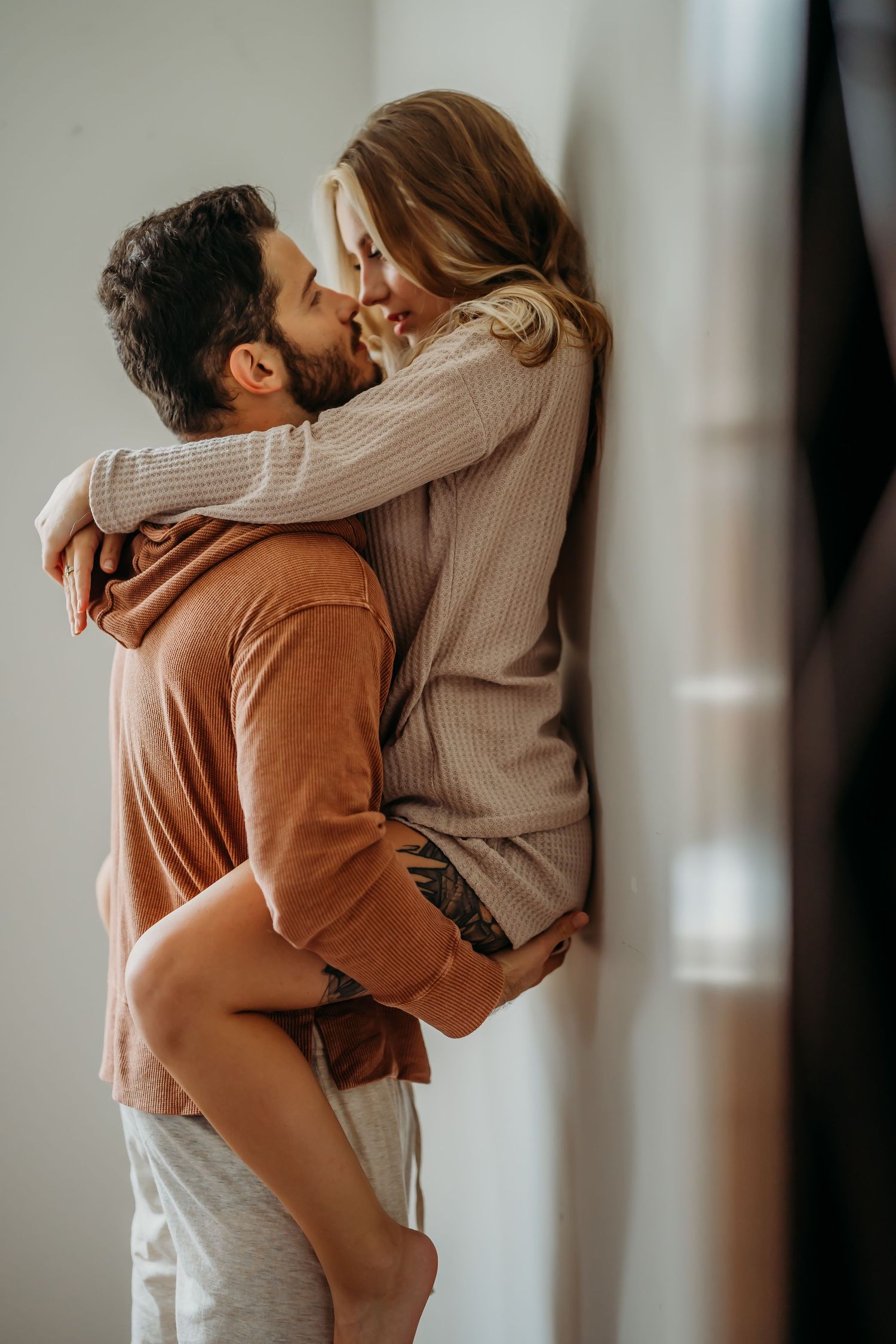 A romantic couple embracing and smiling at each other, the man in an orange hoodie holding the woman in a beige sweater.