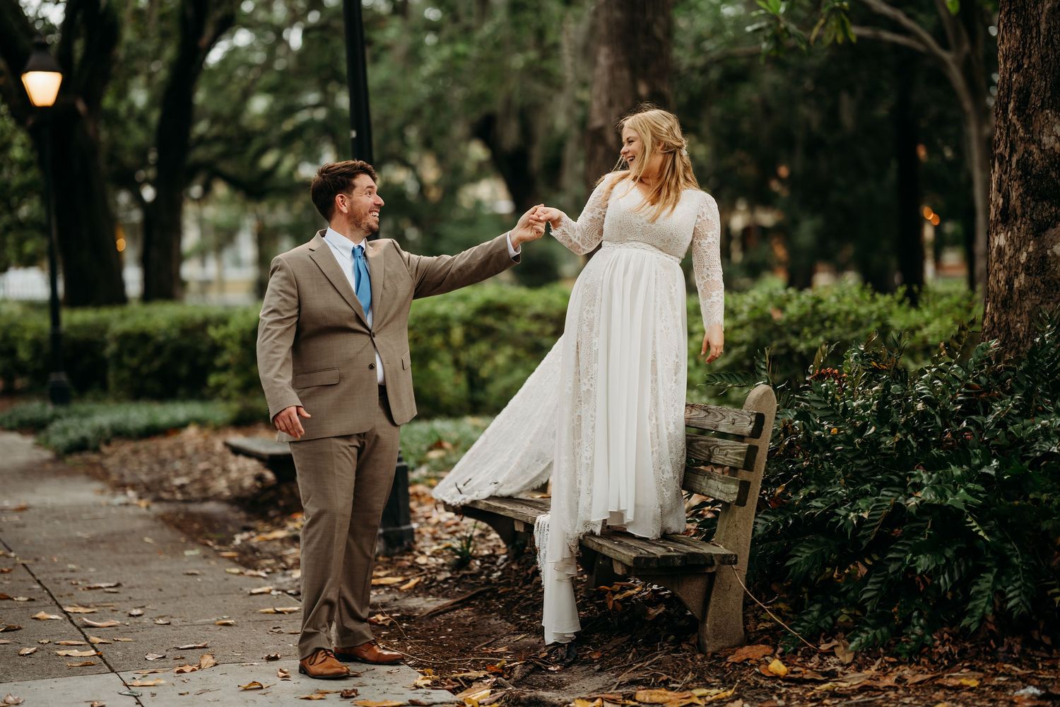 Groom spins bride off a park bench while holding hands on a tree-lined garden path.