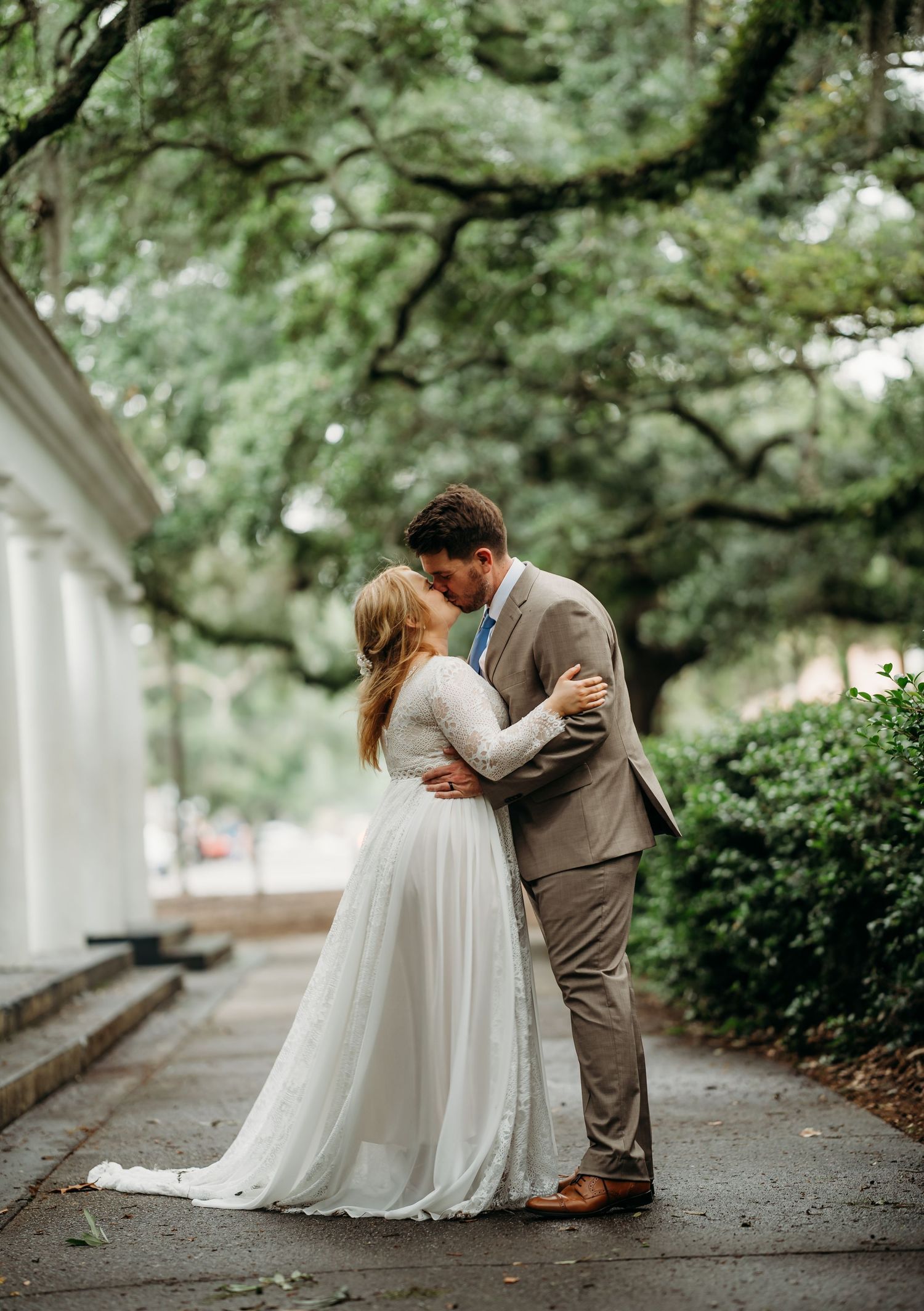 Bride and groom embracing and kissing on a beautiful outdoor pathway lined with white columns and oak trees.