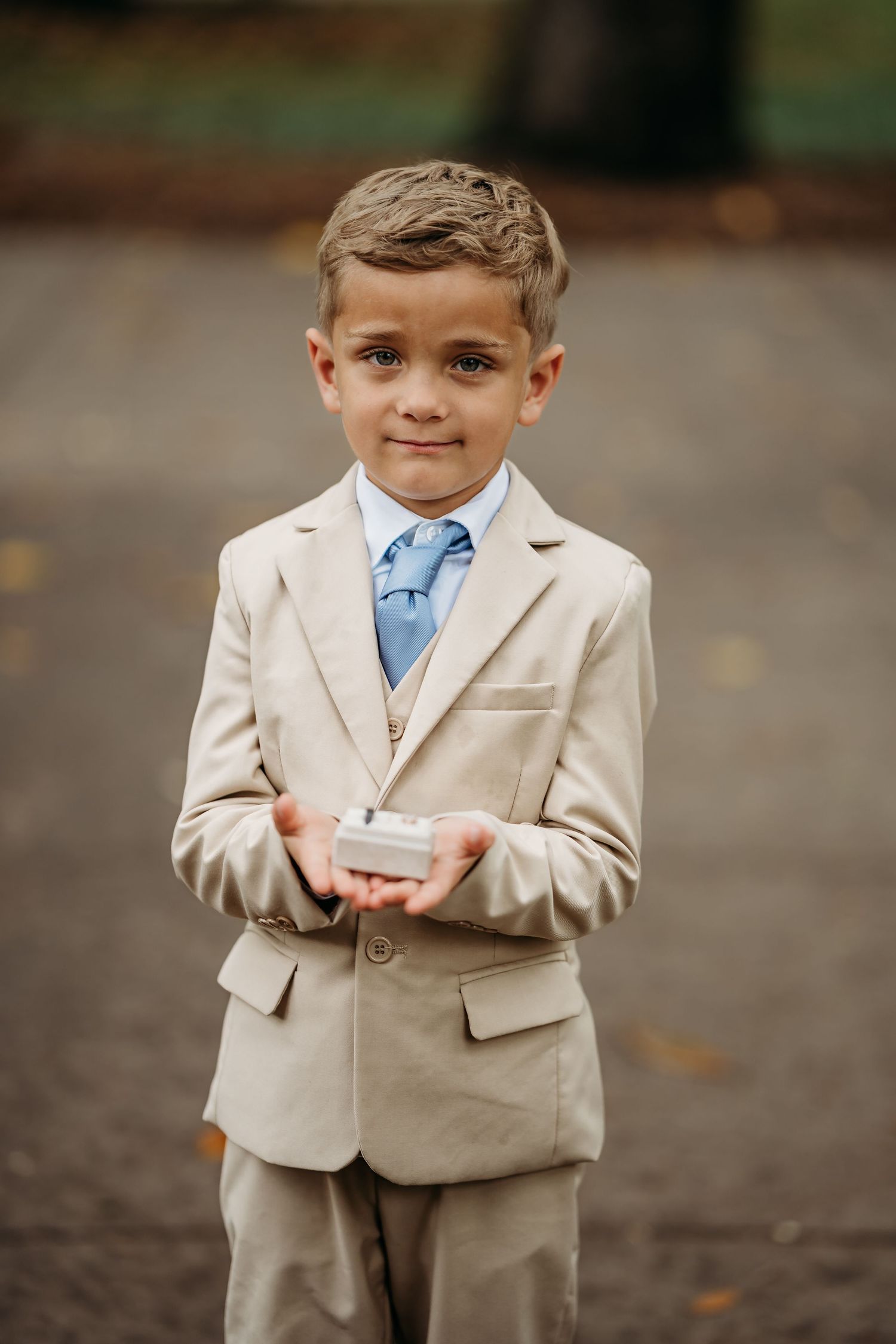 Young ring bearer in beige suit and blue tie holding ring box at wedding.