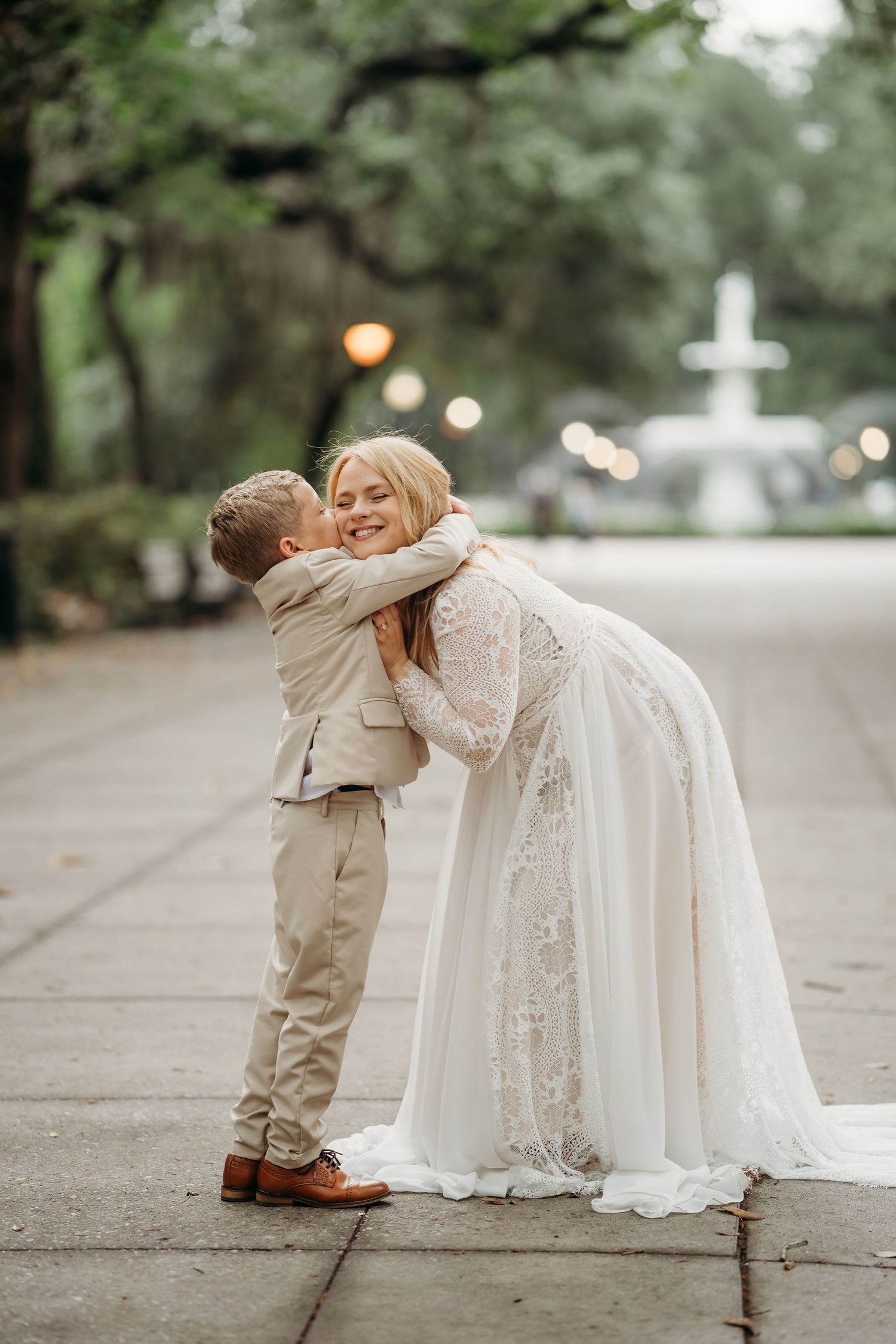 Bride hugging smiling young boy in tan suit on park pathway with fountain backdrop.