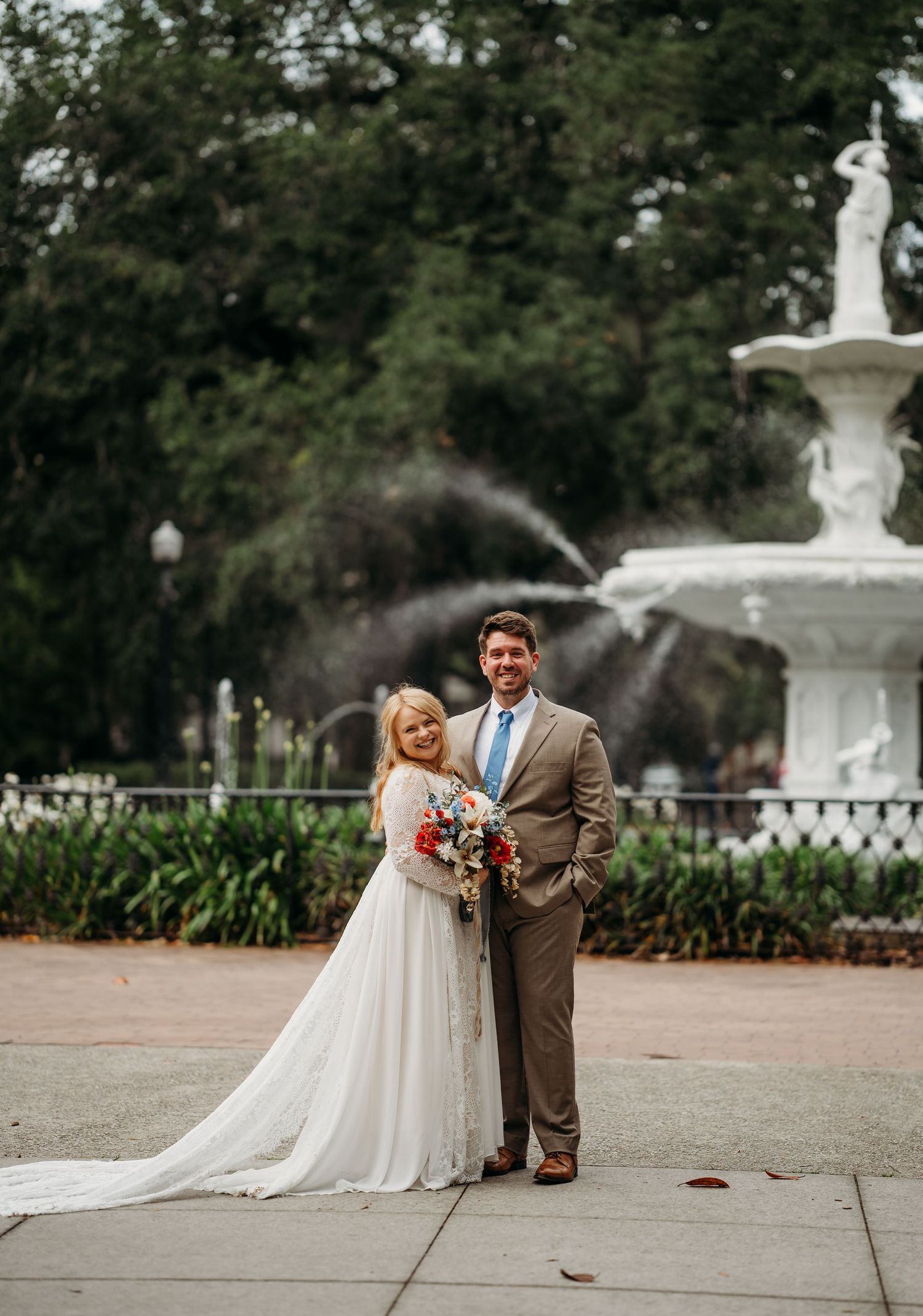 Smiling bride and groom pose together in front of a white fountain, bride holding colorful bouquet.