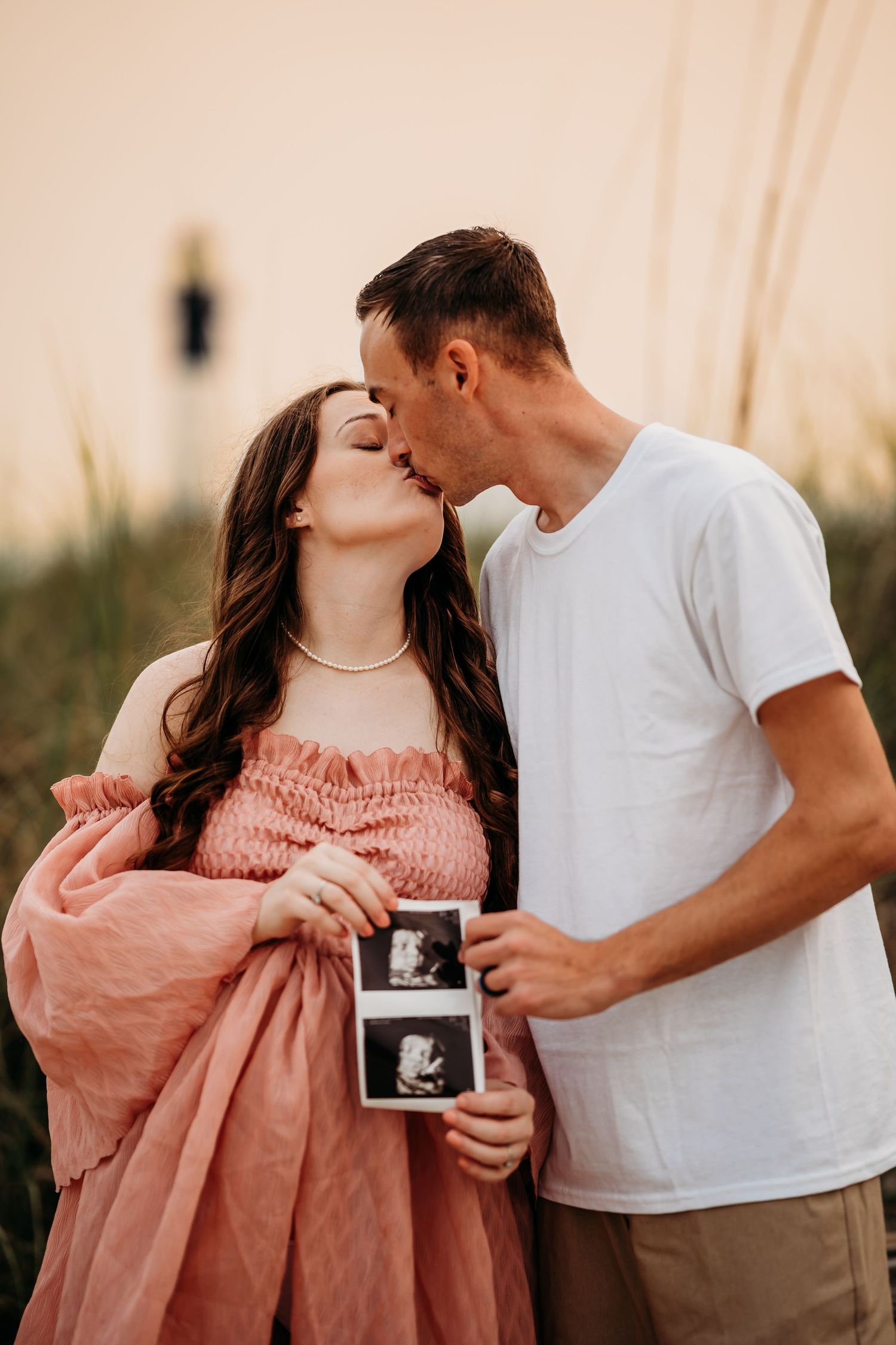 Expecting couple kissing at sunset while holding ultrasound photos during beach maternity shoot.