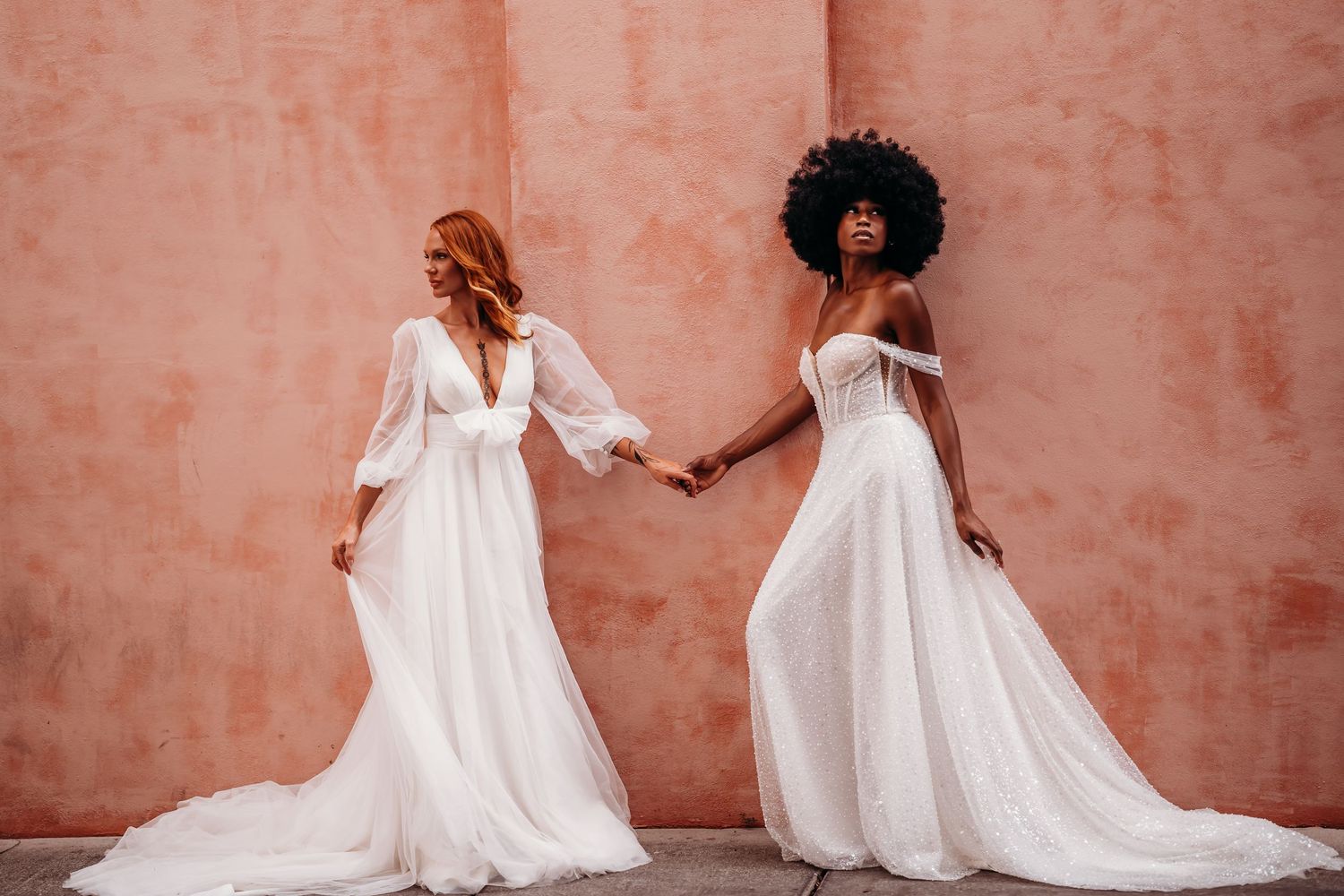 Two brides in elegant white wedding gowns hold hands against a terracotta pink wall.