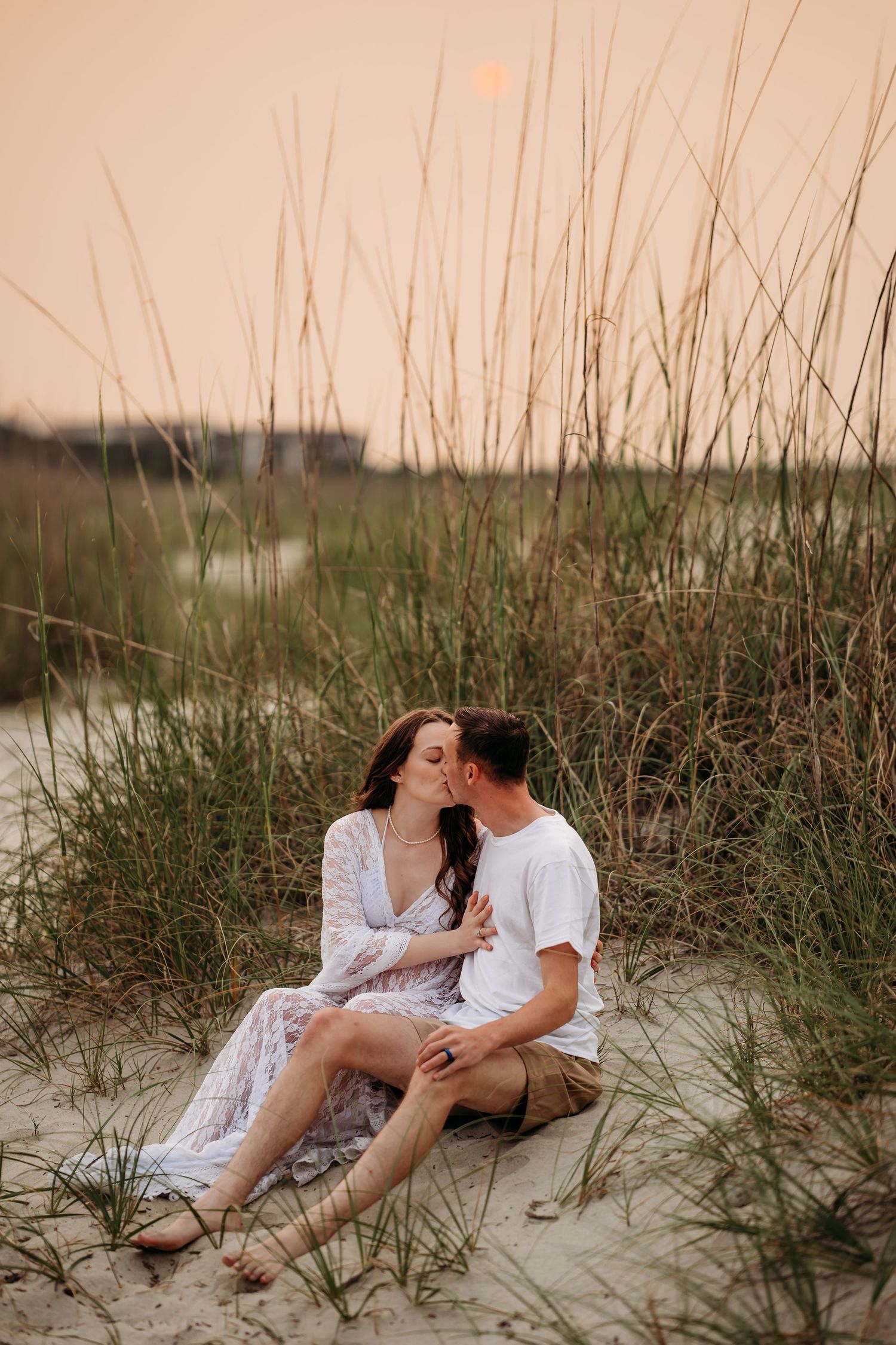 A couple shares a romantic kiss while sitting together in beach grass dunes at golden sunset.