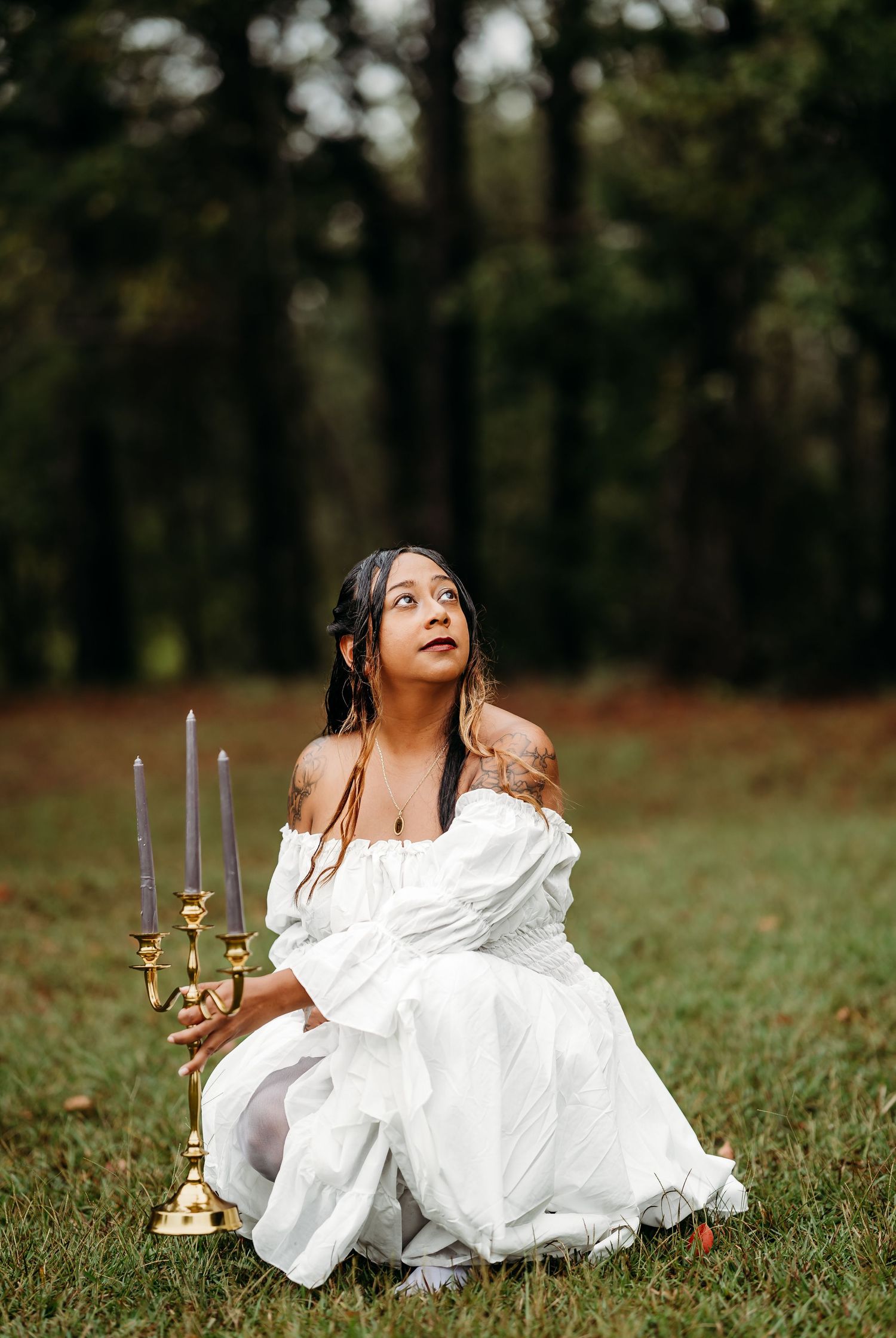 Woman in white off-shoulder dress sits on grass holding gold candelabra with dark candles in forest setting.