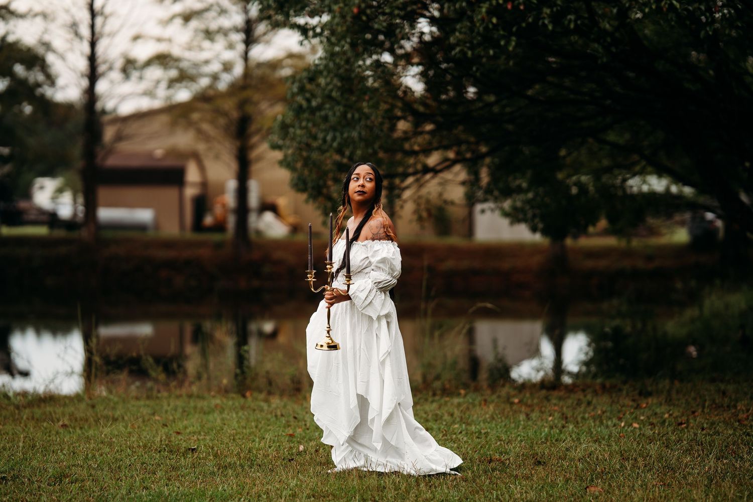 Woman in white off-shoulder gown poses near pond holding gold candelabra in lush outdoor setting.