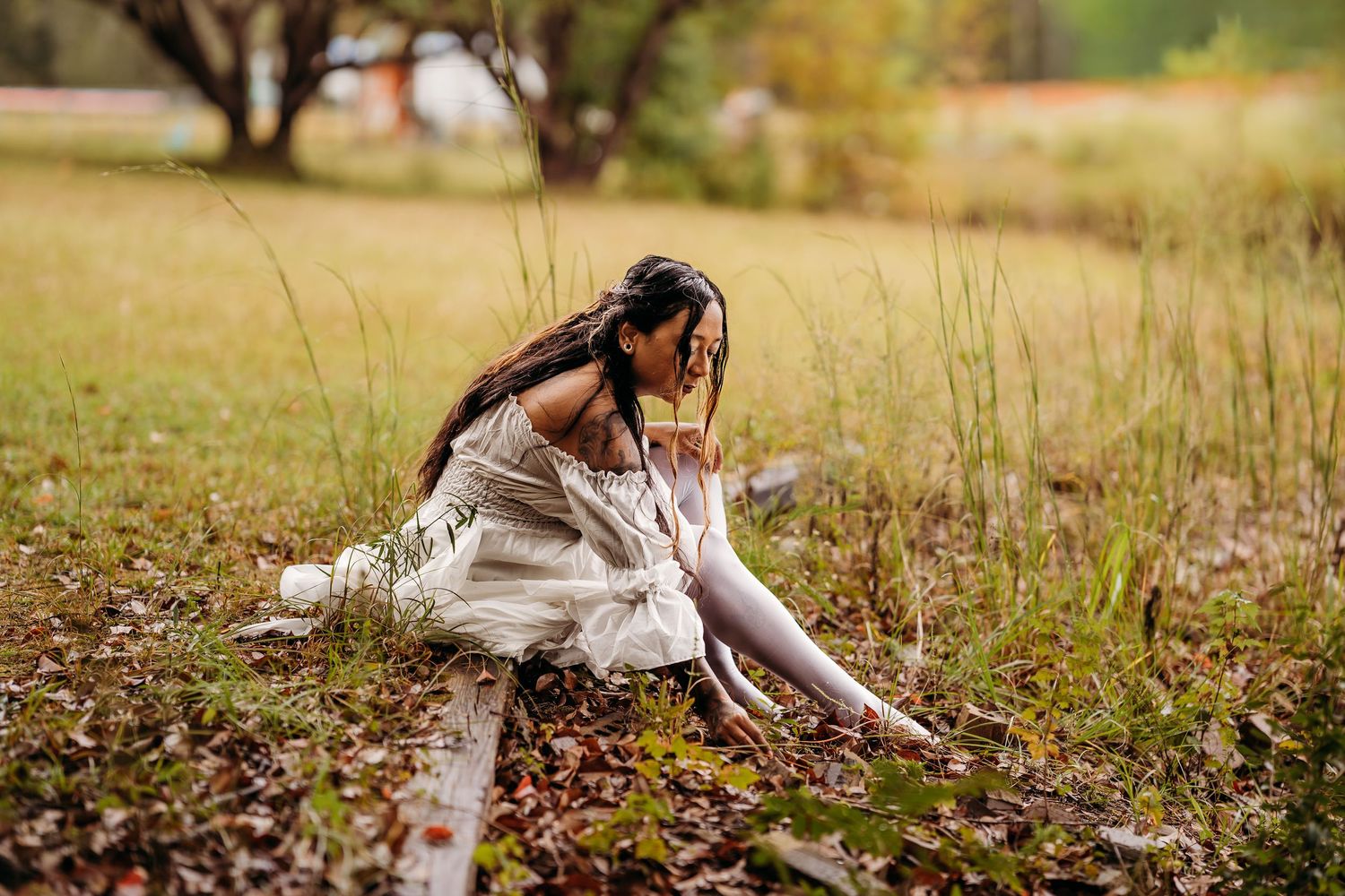 Woman in white dress and white tights sitting on fallen leaves in an autumn field.