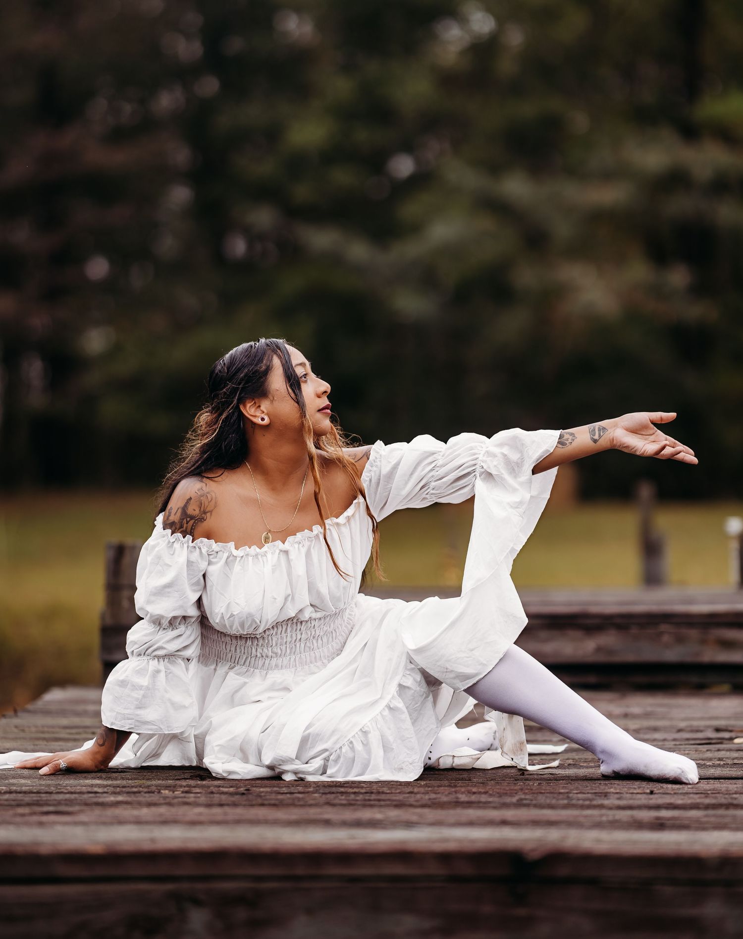 Woman in white off-shoulder dress poses gracefully seated on a wooden dock outdoors.