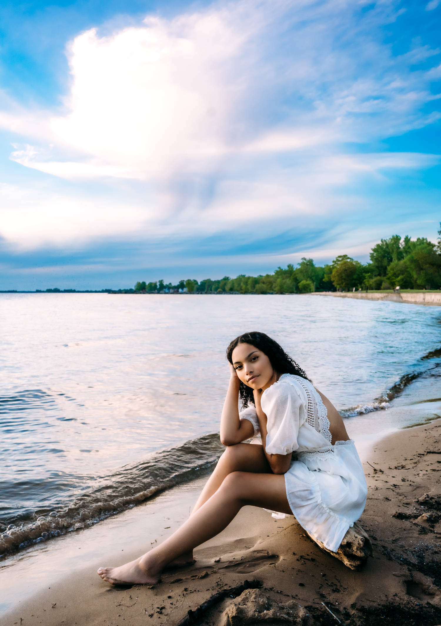 Young woman in white dress sits barefoot on sandy beach shore at sunset with calm water behind her.